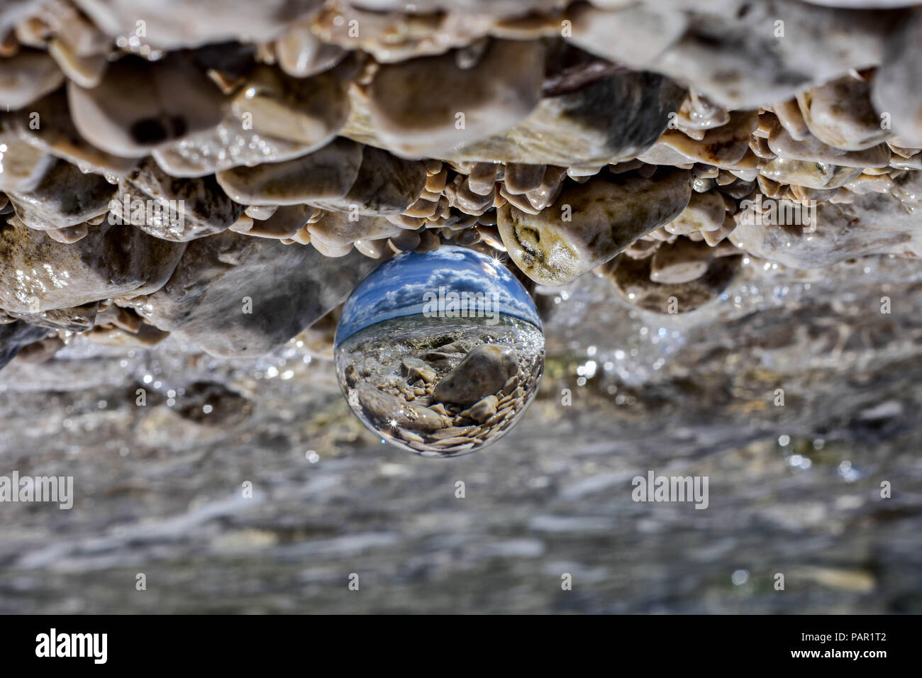Glass ball sitting at rocky beach whit sea wave reflected in it/ conceptual image of summer vacation Stock Photo