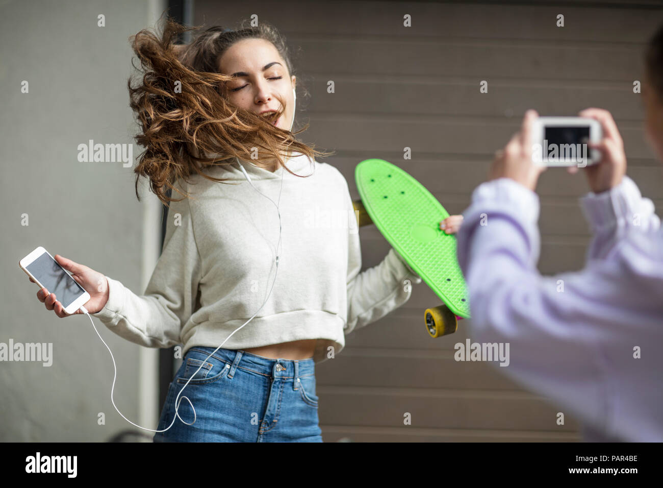 Friend taking picture of carefree teenage girl dancing while holding skateboard and listening to music Stock Photo