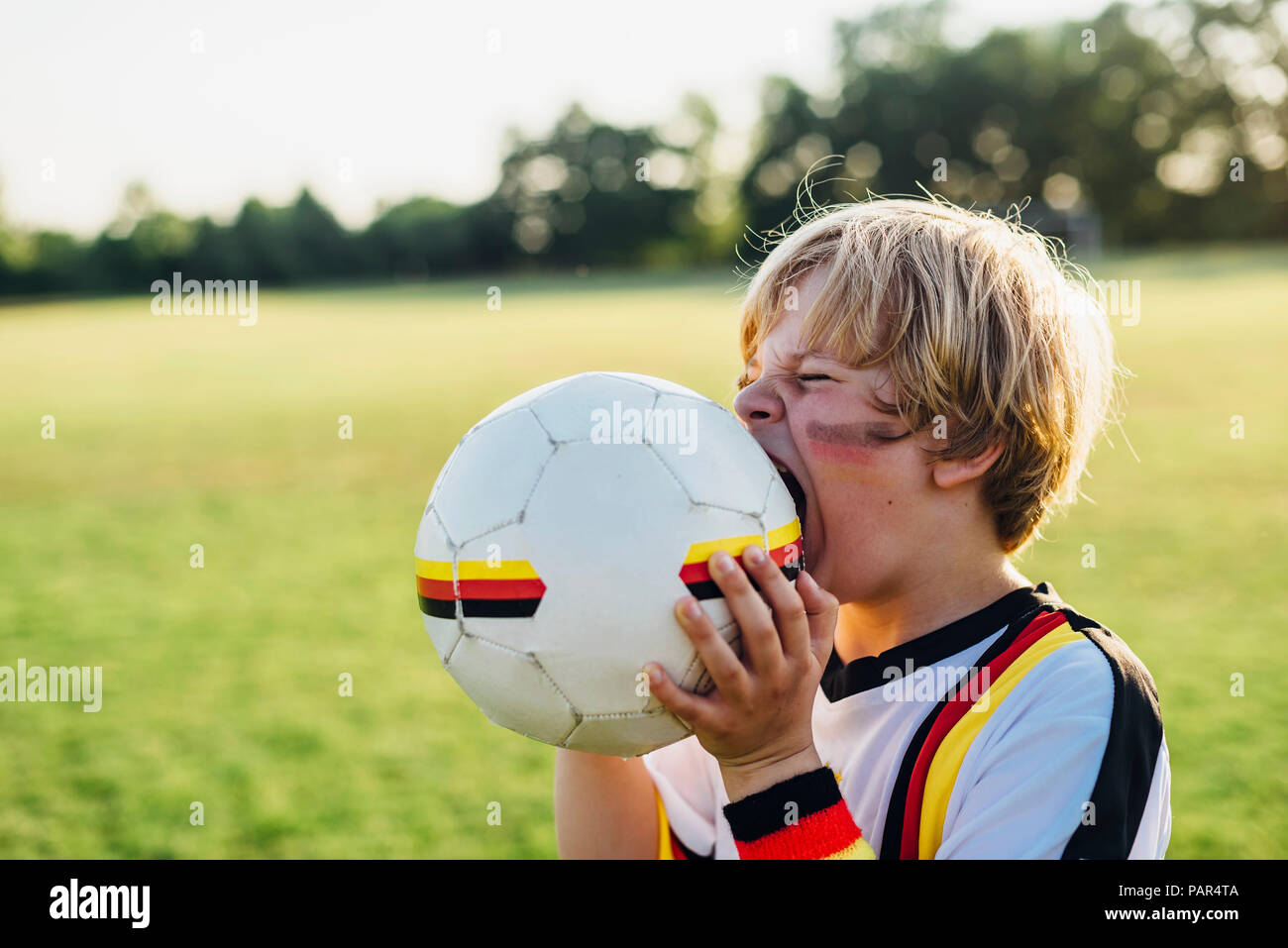 Boy with face paint and German football shirt, biting soccer ball Stock Photo