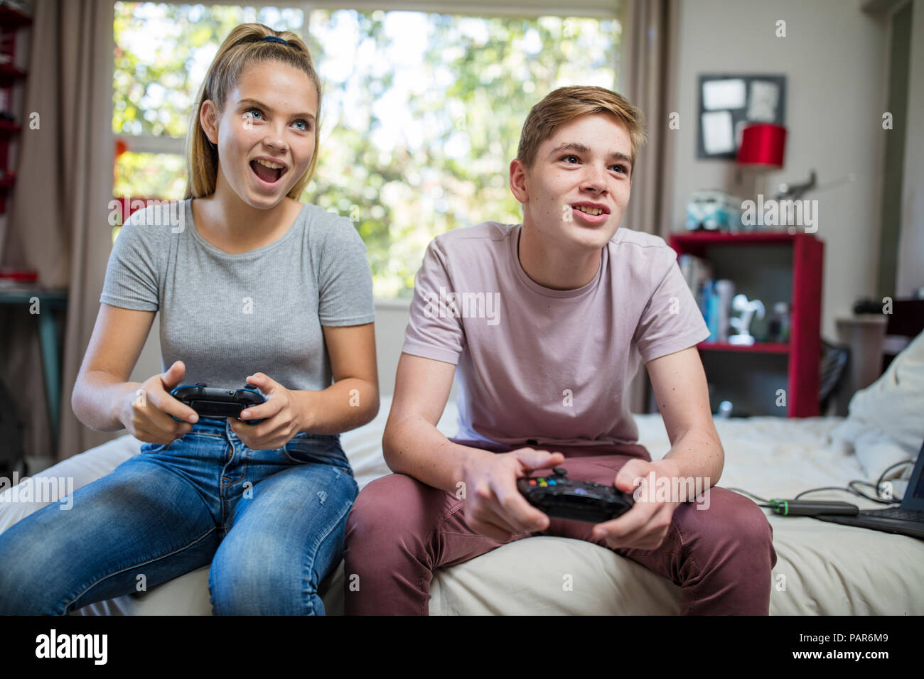 Happy teenage girl and boy sitting on bed playing video game Stock Photo