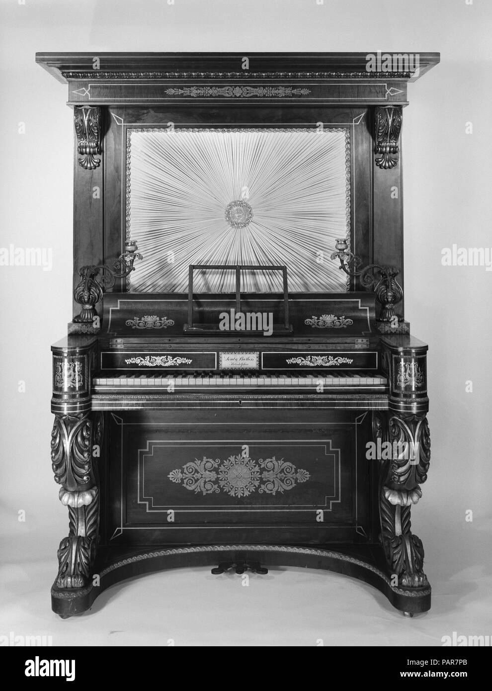 Upright Piano. Culture: American. Dimensions: 76 1/8 × 49 15/16 × 27 15/16 in. (193.3 × 126.9 × 71 cm). Maker: Loud & Brothers (American). Date: 1831.  This large upright piano takes the form of a secretary desk and has a case of carved rosewood with gild decoration. Its fabric panel (a modern replacement) the piano action. The keyboard compass is 73 keys (FF-f4) with ivory naturals and ebony accidentals. Two brass pivoting candleholders flank the keyboard.  There are three wooden pedals, the right raises the dampers, the center shifts the action for an una corda, and the left opens swell shut Stock Photo