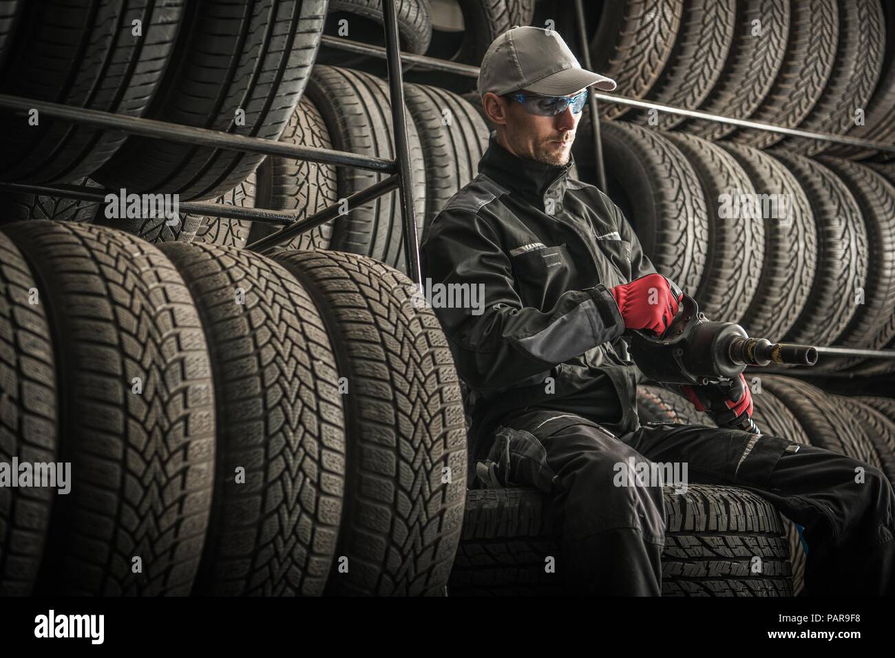 Car Tires Sales and Repair. Service Worker with Power Tool Between New and Used Tires. Automotive Theme. Seasonal Vehicle Tires Changes. Stock Photo