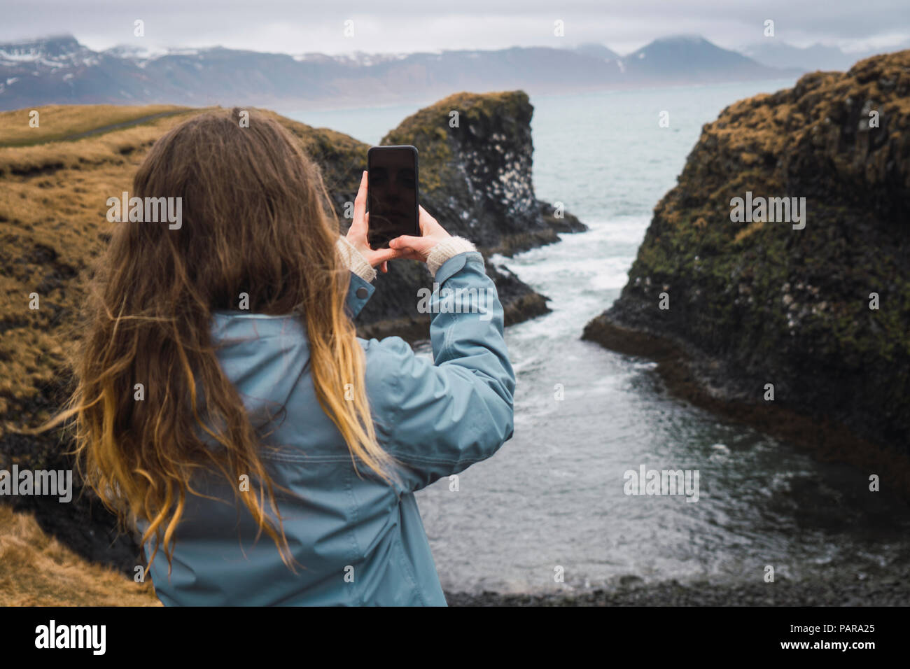 Iceland, back view of young woman taking picture with smartphone at coast Stock Photo