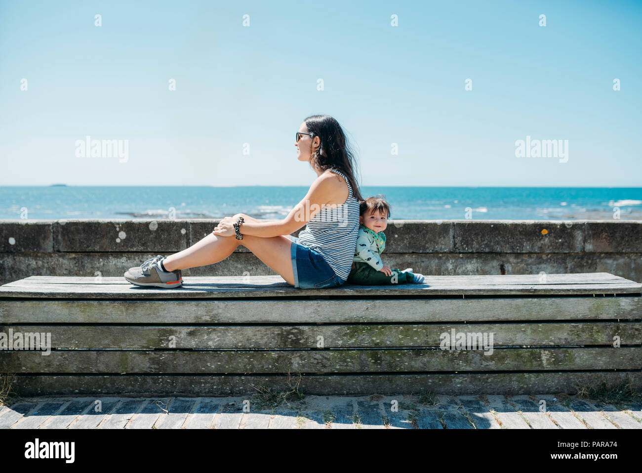 France, mother and baby girl sitting back to back on a bench at beach promenade Stock Photo