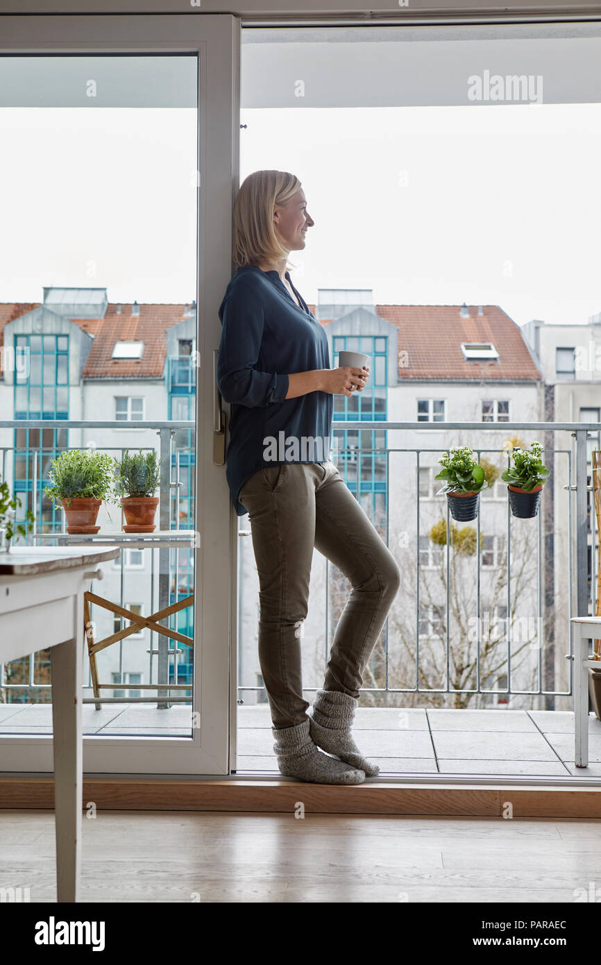 Smiling woman holding cup of coffee looking out of balcony door Stock Photo