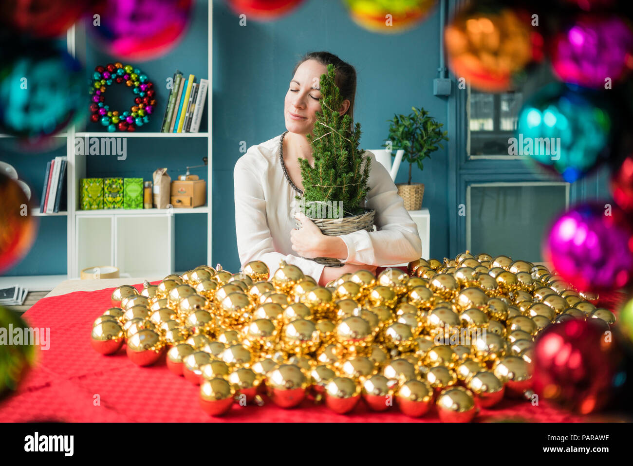 Woman sitting at table with many golden Christmas baubles holding potted fir tree Stock Photo