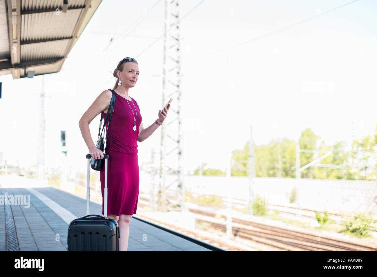 Mature businesswoman with suitcase and smartphone waiting at platform Stock Photo
