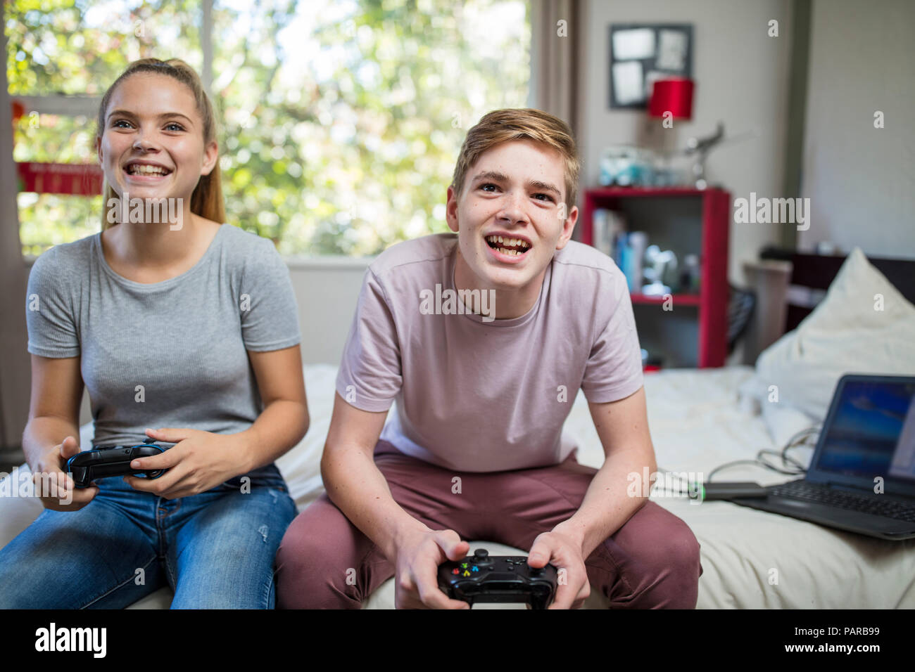 Happy teenage girl and boy sitting on bed playing video game Stock Photo