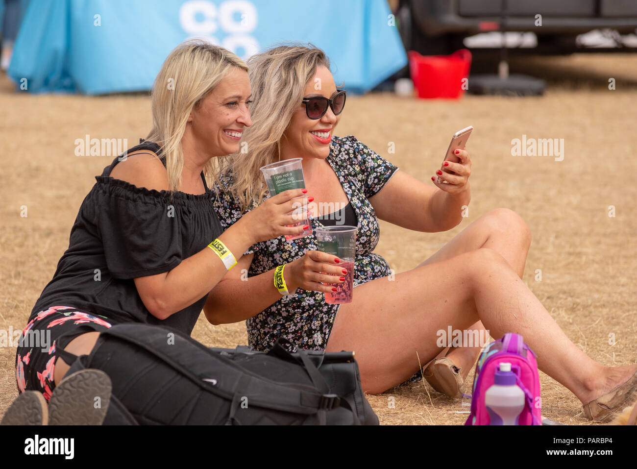 Two attractive blond women sitting down smiling and taking a selfie at a summer festival while holding drinks. Stock Photo