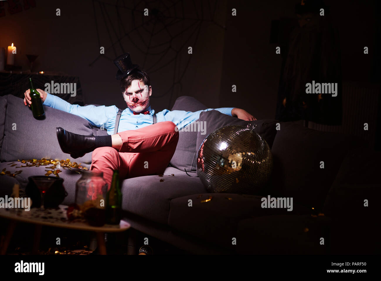 Man in Halloween costume sitting on couch after party, drinking beer Stock Photo