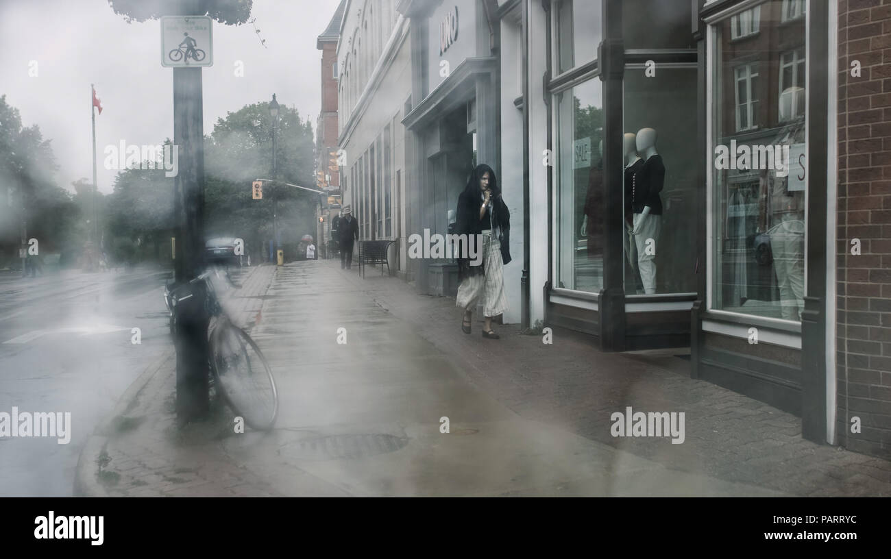 It's raining in Stratford, a woman, decidedly in a hurry, protects her head and walks fast, while throwing a glance at a shop window. Stock Photo