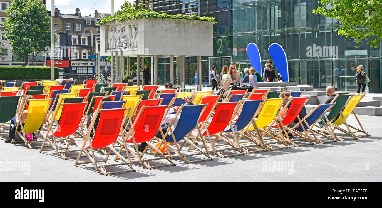 People enjoying sitting in summer sunshine colourful deckchairs outside office block near coffee & food bars just before lunch time London England UK Stock Photo