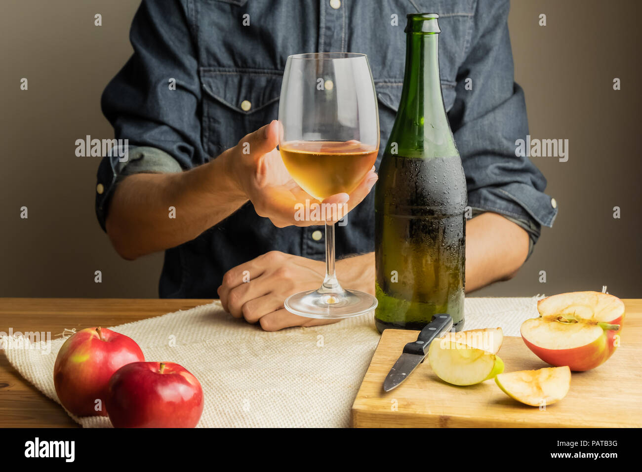 Beautiful ice cold glass and bottle of apple wine, with ripe apples in background. Male hands holding glass of premium cider on rustic wood table. Stock Photo