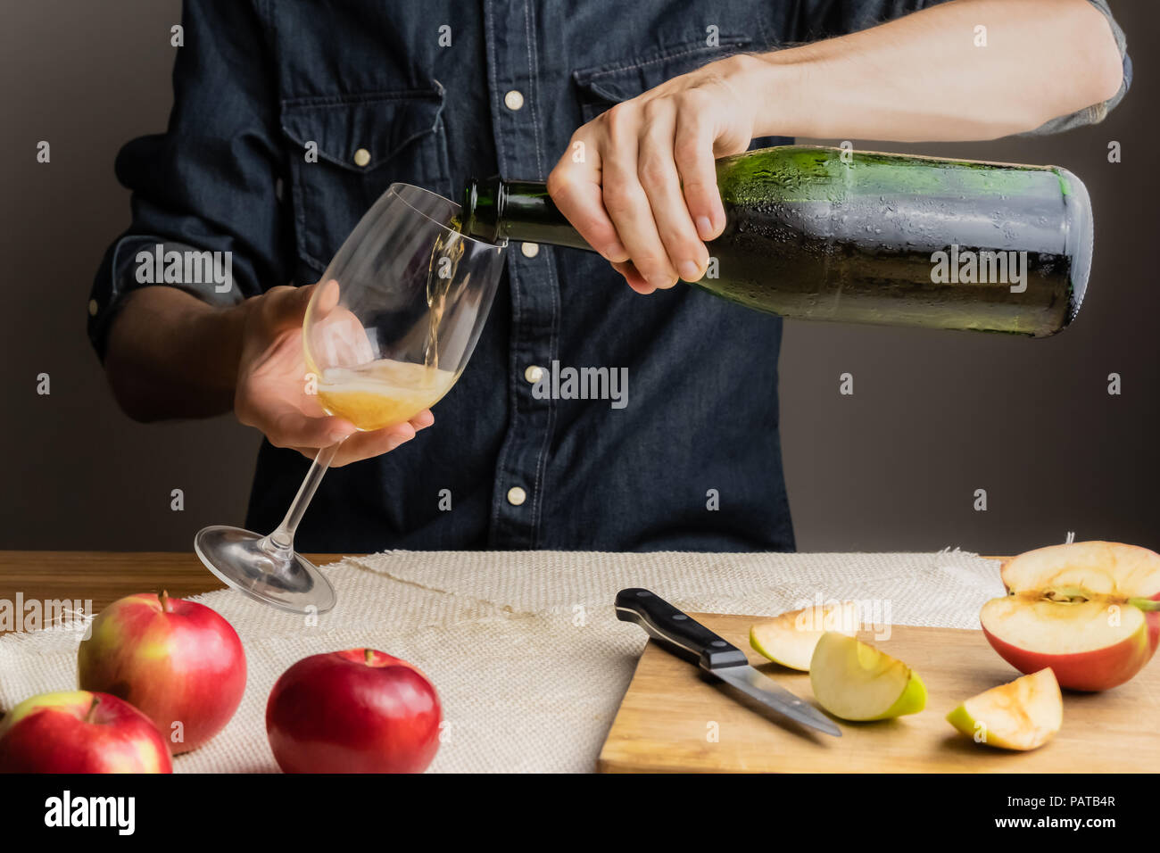 Man pours a glass of vintage apple wine out of ice cold bottle. Male hands pouring premium cidre in wine glass above rustic wood table. Stock Photo