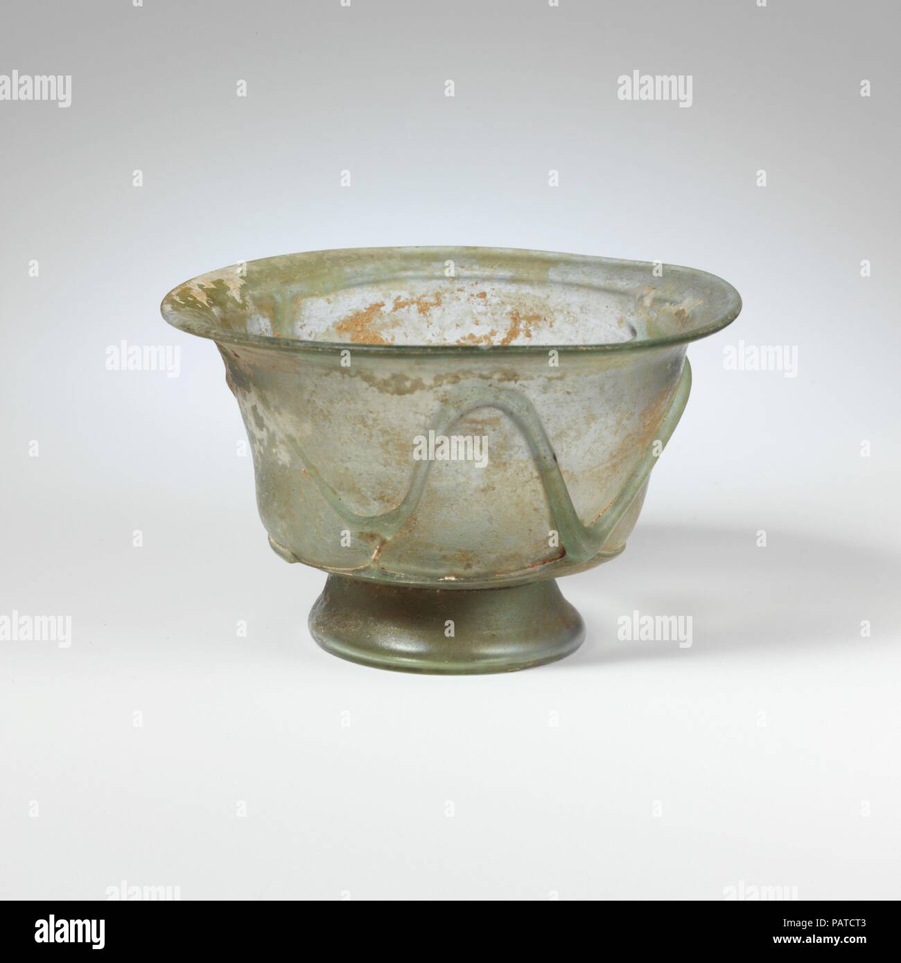 Glass bowl. Culture: Roman. Dimensions: Overall: 2 5/8in. (6.7cm)  Other: 4 1/16in. (10.3cm). Date: 4th century A.D..  Translucent blue green; trails in same color.  Outsplayed rim, with rounded lip; side of body tapers downwards, then curves inwards; tall, splayed tubular foot, made by folding; thick, conical bottom, flattened at center by pontil mark.  A fine trail applied horizontally around base of side; another thicker trail applied over it and wound round side in an irregular, curving zigzag pattern, ending in a long trail under rim.  Intact; pinprick bubbles and blowing striations, with Stock Photo
