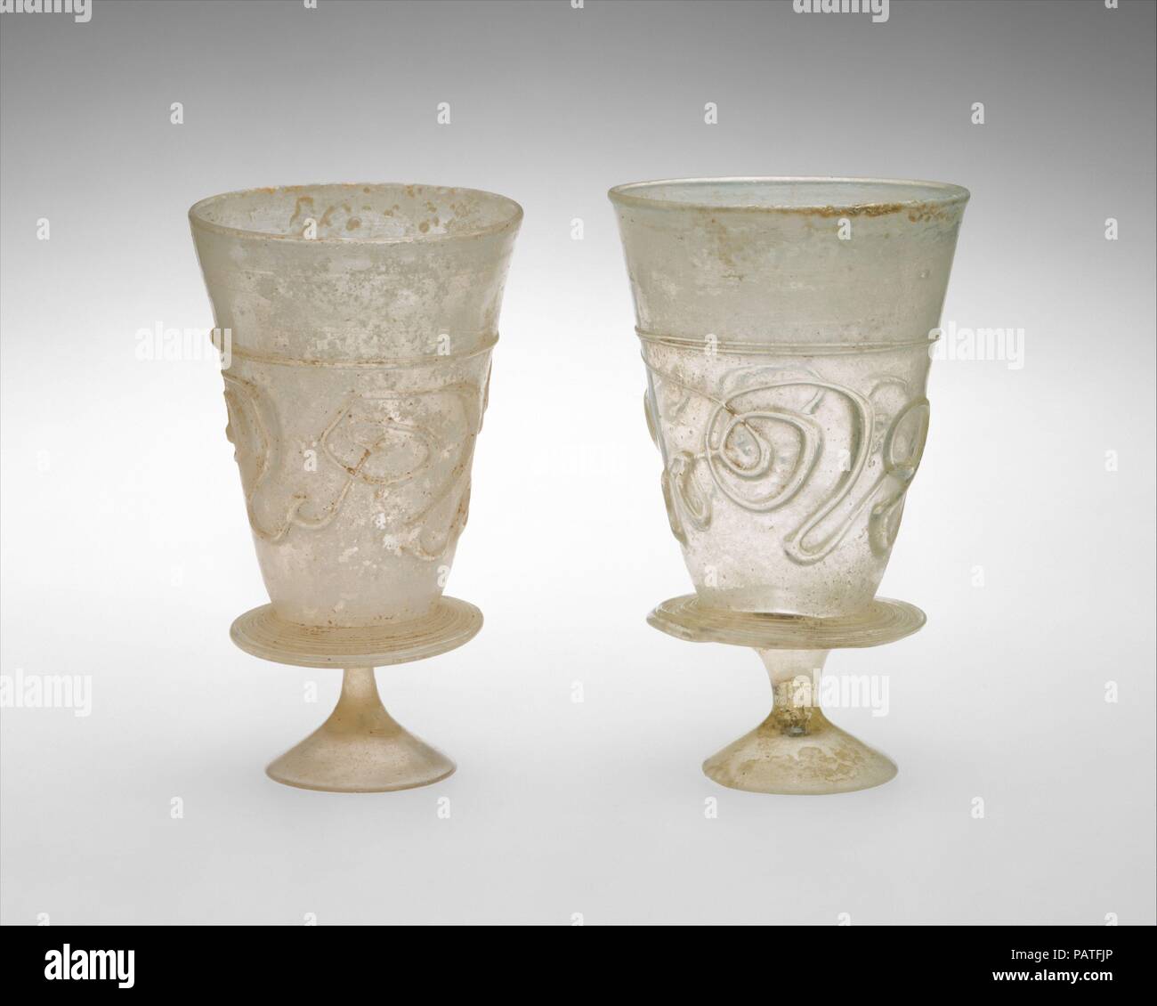 Goblets with Applied Decoration. Date: 11th-early 12th century.  These matcing goblets are each formed by a conical cup attached to a small, solid, and splayed stemmed foot by a circular flange applied around the base of the cup. They are made of yellowish colorless glass that contains many small bubbles. Both cups are decorated with an unbroken applied trail in the same yellowish color, which forms a horizontal line about two-thirds of the height and continues below to create a fanciful, abstract pattern of curly designs around the cup. The decoration can be read more clearly when the cup is  Stock Photo