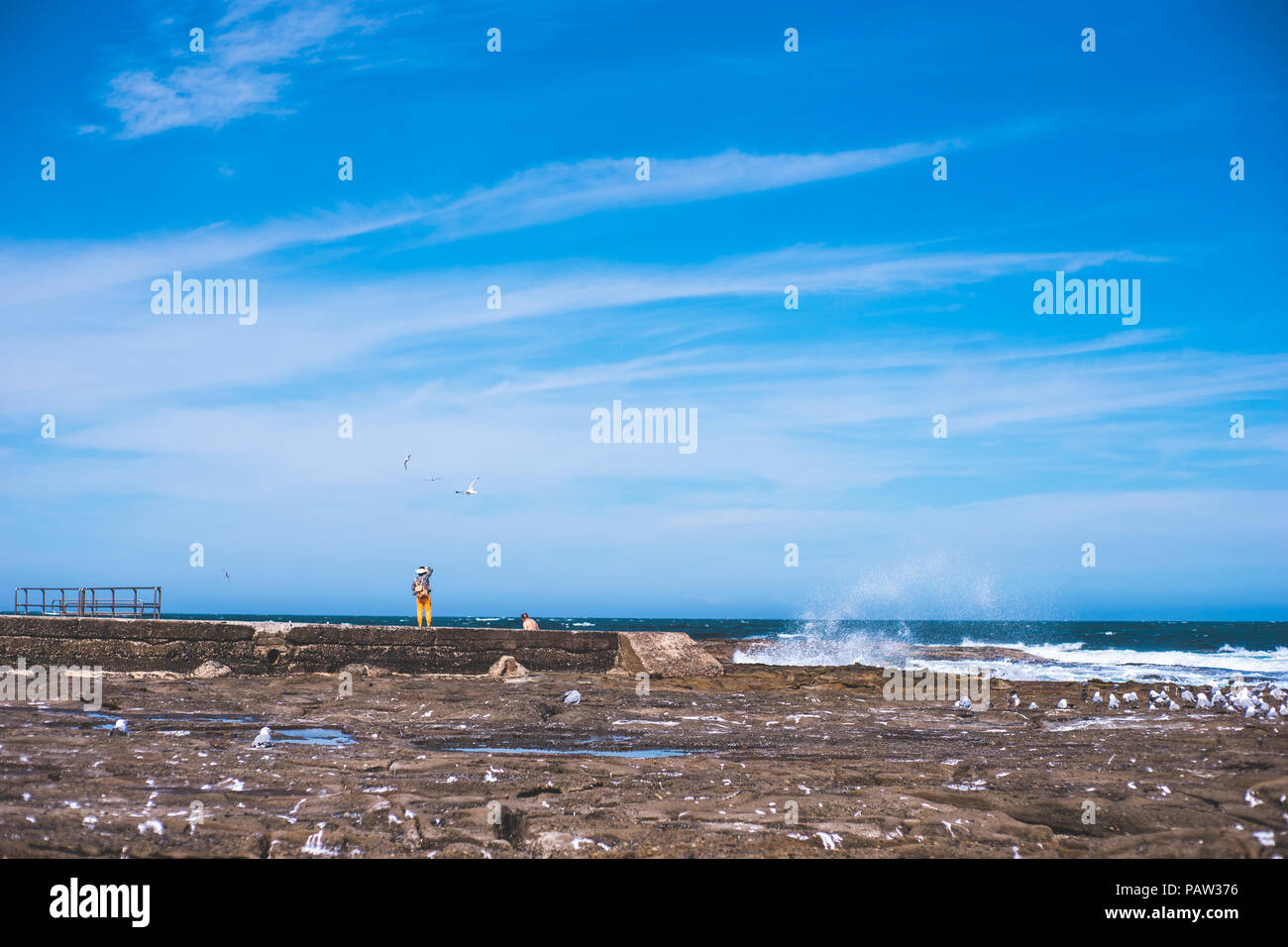 minimal scenery of blue sky and rock beach with a tiny lady standing on the edge of ocean bath in suuny day Stock Photo