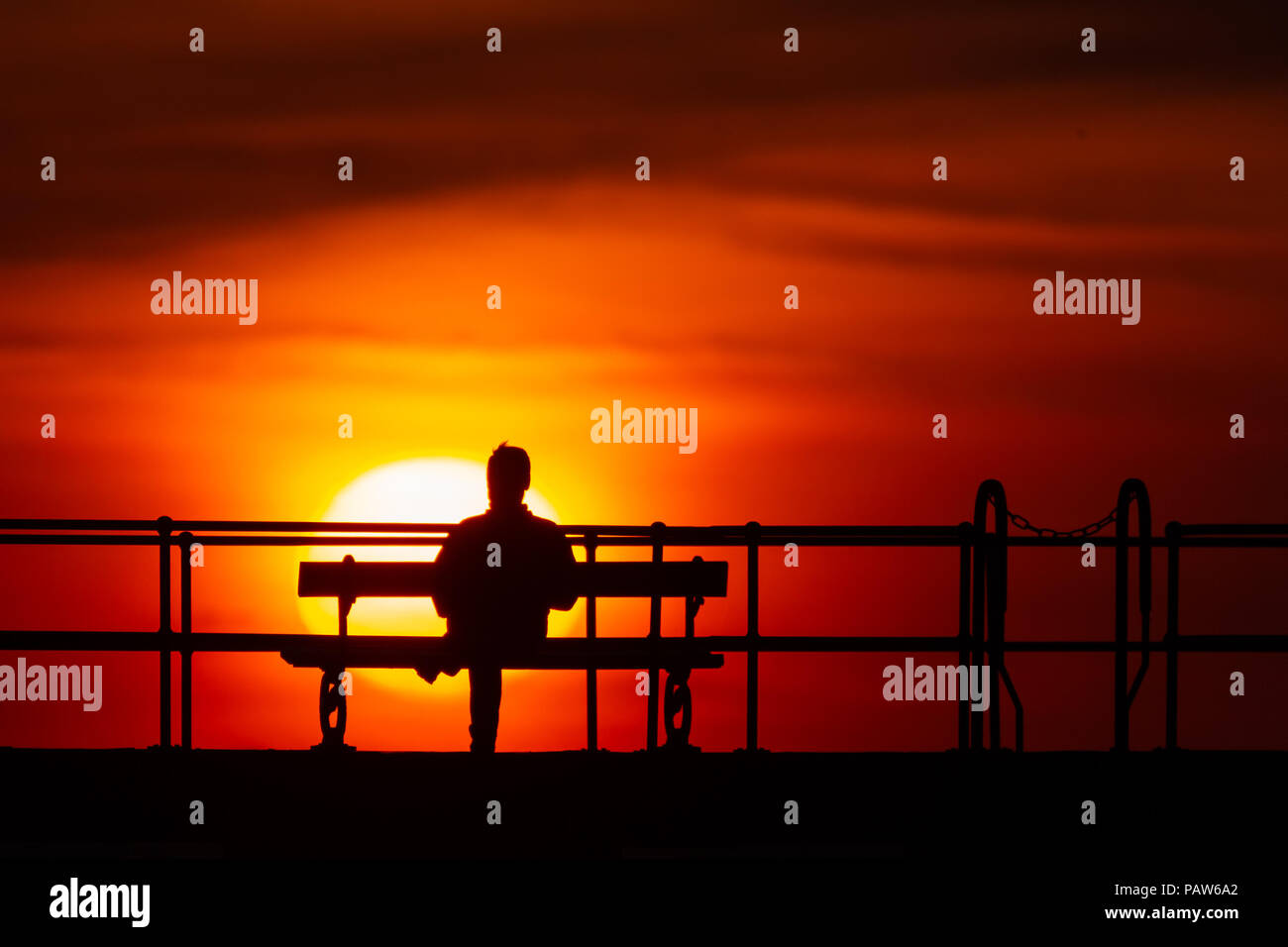 Silhouetted man sitting on a bench along a railed walkway, watching the sun go down at sunset. © Ian Jones/Alamy Live News. Stock Photo