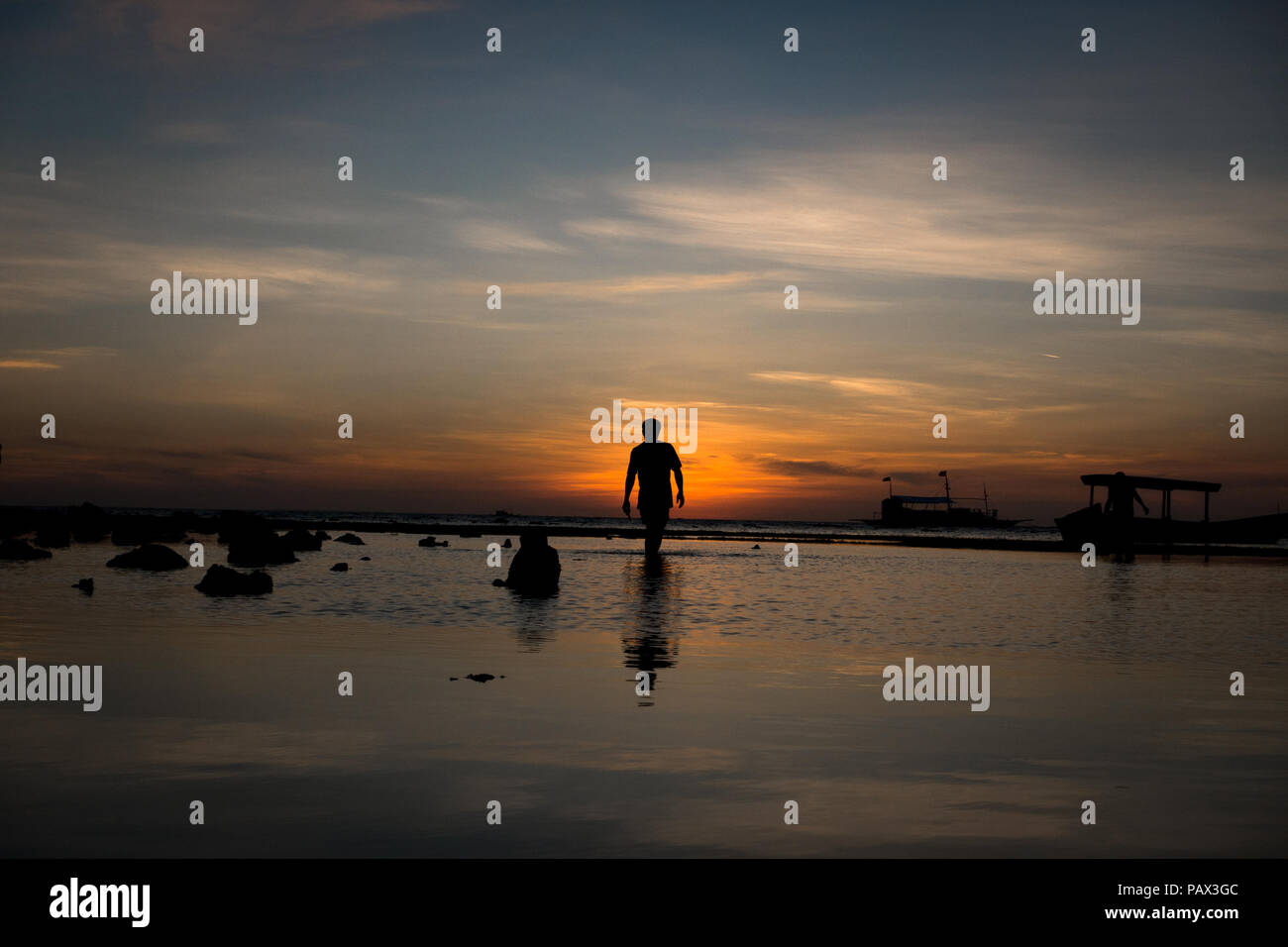 Silhouette of a lone man walking along the water's edge during a low tide island sunset - Malapascua Cebu - Philippines Stock Photo