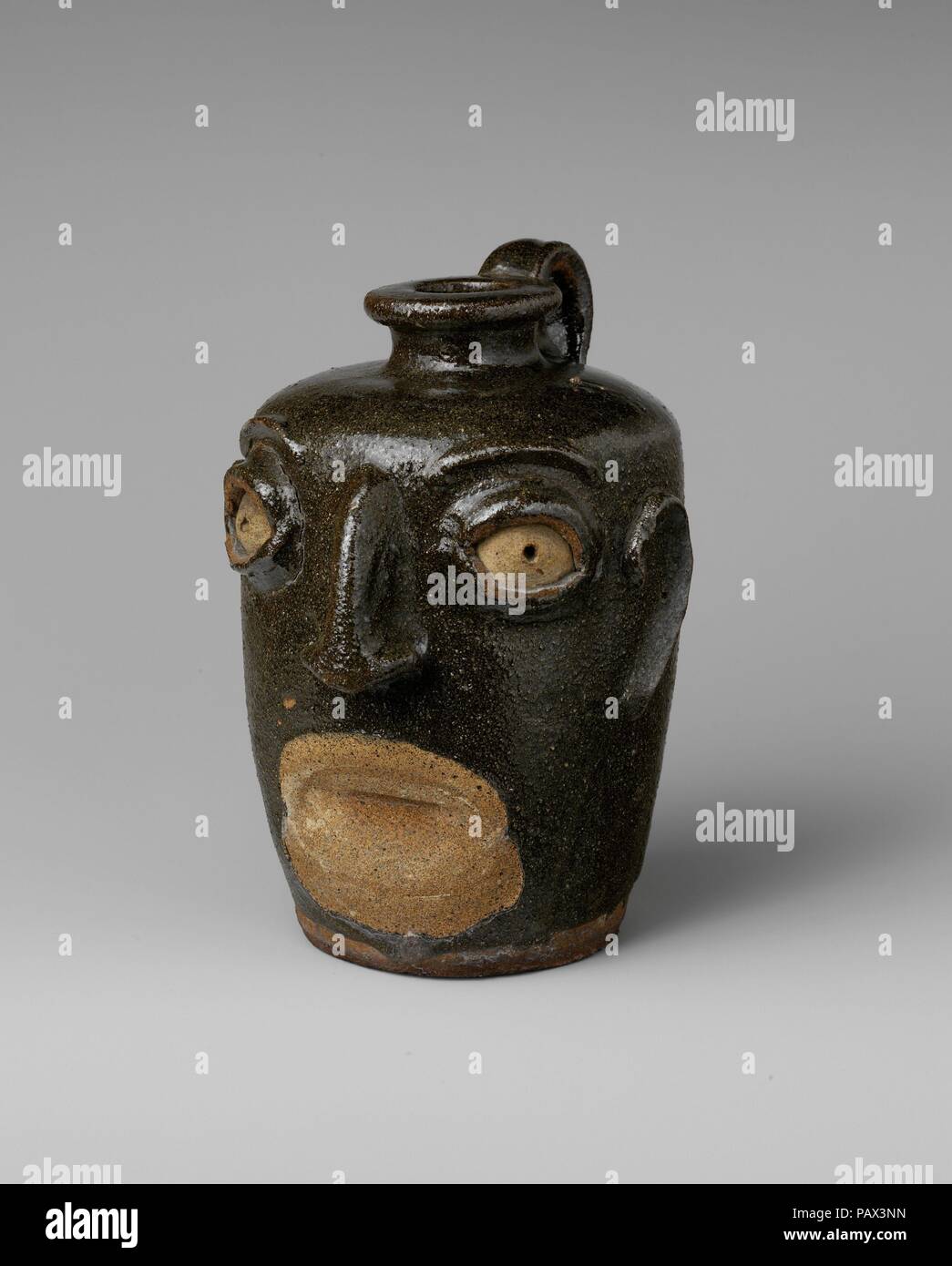 Face Jug. Culture: American. Dimensions: 7 x 5 x 5 1/2 in. (17.8 x 12.7 x 14 cm). Maker: John Lewis Miles Pottery (1868-75). Date: ca. 1860-70.  Face jugs were made by African American slaves and freedmen working in potteries in the Edgefield District of South Carolina, an area of significant stoneware production in the nineteenth century. The distinctive features of the jugs, notably the kaolin inserts for the eyes, relate in style and material to ritualistic objects of the Congo and Angola region of western Africa, whence many slaves in South Carolina descended. This jug is missing its teeth Stock Photo