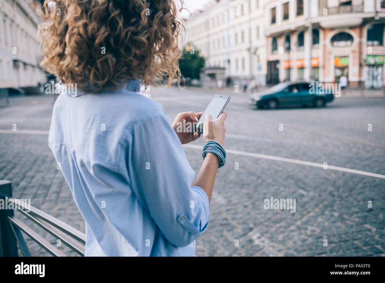 Rear view unrecognizable curly young woman using smart phone standing near european paving stone road with cars. Female wearing blue shirt and bracele Stock Photo