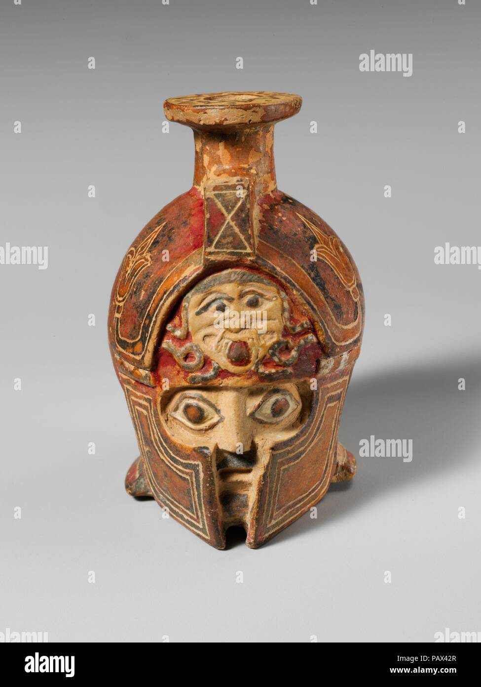 Terracotta aryballos in the form of a helmeted head. Culture: East Greek. Dimensions: H. 7.01 cm.. Date: ca. 600-575 B.C..  A helmeted warrior's head makes up the body of this small mold-made terracotta aryballos, likely used to hold perfumed oil. The painted and incised surface is very well preserved, including a prominent gorgoneion on the metopon of the Ionian helmet. The warrior's wide eyes and dark beard are visible through the gaps in the helmet. Museum: Metropolitan Museum of Art, New York, USA. Stock Photo