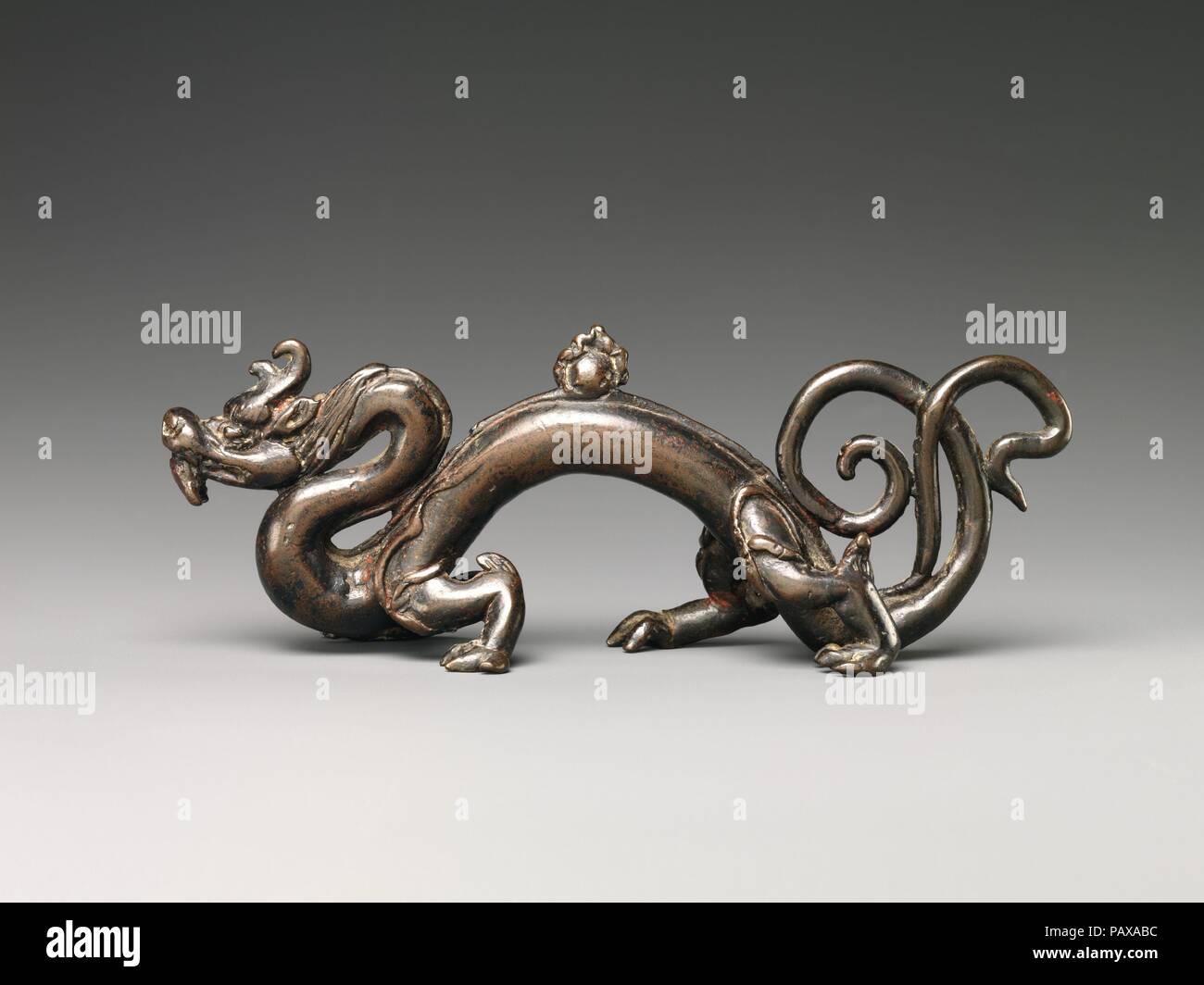 Brush Rest in the Form of a Dragon. Culture: China. Dimensions: H. 2 1/8 in. (5.4 cm); W. 6 7/8 in. (17.5 cm); D. 1 1/2 in. (3.8 cm). Date: 15th century.  A rare example of 15th century Ming bronze work, this brush rest was cast in the form of a sinuous dragon crouching on its four powerful paws with a 'flaming pearl' on its arched back. It is further distinguished by two curved horns and a bifurcated tail that coils and unfurls. Its neck, body, and tail all provide places for resting writing brushes. More than a functional object, this brush rest is an imaginative and auspicious sculpture des Stock Photo