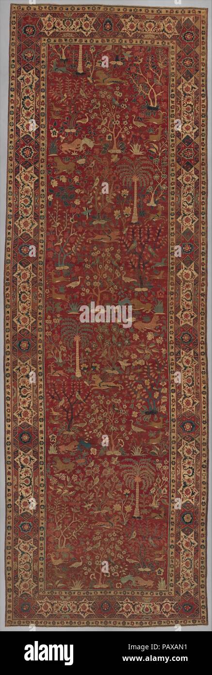 Carpet with Palm Trees, Ibexes, and Birds. Dimensions: Rug: L. 328 in. (833.1 cm)  W. 108 in. (274.3 cm)  Wt. 132 lbs. (59.9 kg)  Storage Tube: L. 132 in. (335.3 cm)  Diam. 9 in. (22.9 cm). Date: late 16th-early 17th century.  This carpet, with its pictorial depiction of trees, birds, and animals, is conceived like a textile with a repeat design in which each unit reverses the direction of the preceding one. The ibexes, Chinese mythological beasts called qilins, and animals in combat, are derived from Safavid Persian art, as is the border design of cartouches and star-shaped medallions with cl Stock Photo