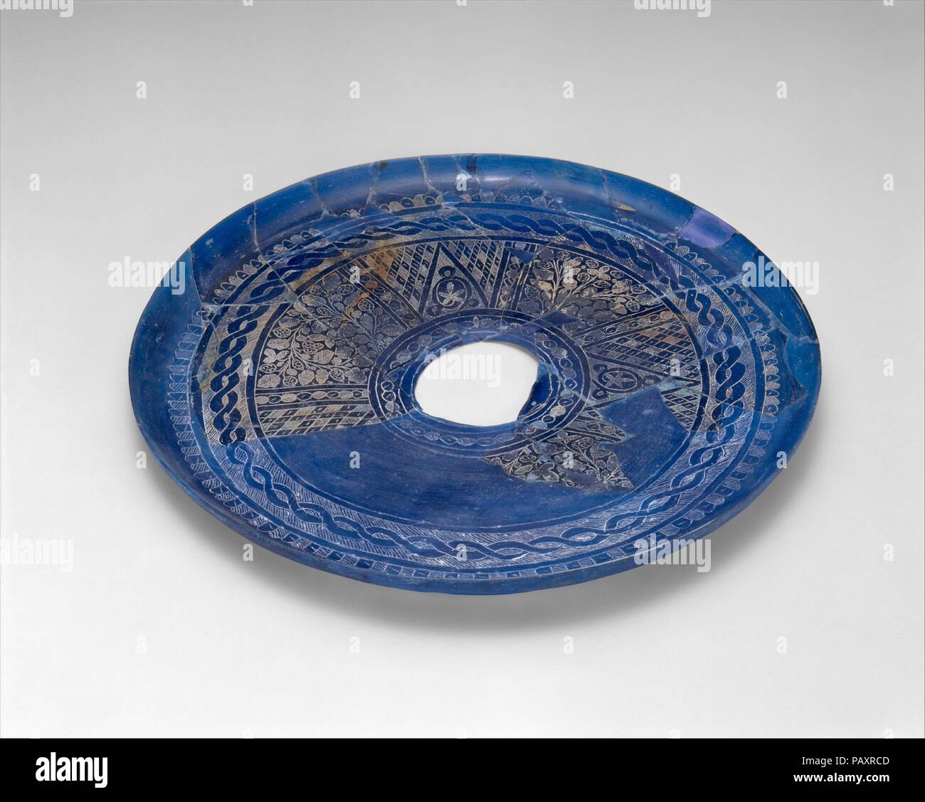 Fragmentary Plate with Engraved Designs. Dimensions: H. ca. 9/16 in. (ca. 1.5 cm)  Diam. 11 in. (28 cm). Date: 9th century.  The striking blue color of this plate was achieved by adding cobalt to the glass fabric, and its surface has been engraved with various patterns arranged in concentric registers around a central roundel. The circular hole at the center indicates the place where a foot was once attached. This plate is important evidence of the active glass trade from west to east during the ninth and tenth centuries. Probably made in Syria, it was imported to Nishapur where it was found i Stock Photo