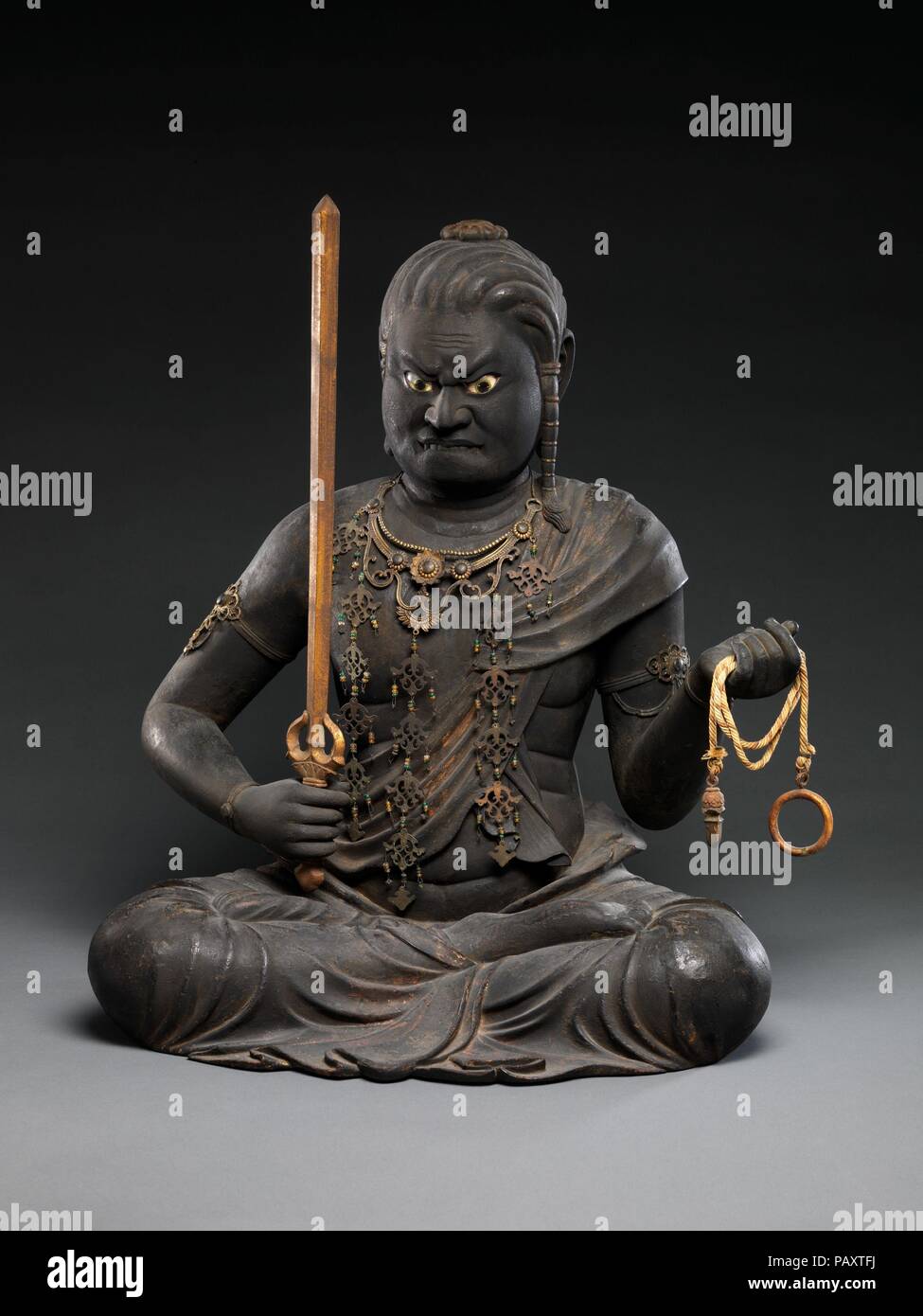Fudo Myoo. Artist: Kaikei (Japanese, active 1183-1223). Culture: Japan. Dimensions: H. 21 in. (53.3 cm); H. to top of sword 21 1/2 in. (54.6 cm); W. 16 3/4 in. (42.5 cm); D. 15 in. (38.1 cm). Date: early 13th century.  Fudo Myoo (Sanskrit: Acala-vidyaraja), the chief of the Five Wisdom Kings (Godai myoo), is the wrathful avatar of Dainichi Buddha and the tenacious protector of Buddhist law. His iconography, drawn from the Dainichi Sutra, describes his body as black or blue, with bulging eyes, protruding fangs that bite his lower lip, and hair that hangs down his left shoulder. He carries in hi Stock Photo