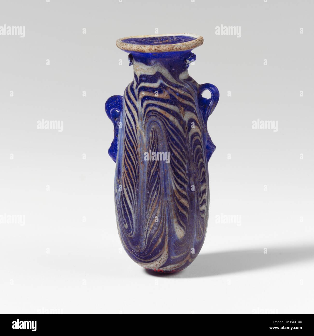 Glass alabastron (perfume bottle). Culture: Greek, Eastern Mediterranean. Dimensions: H.: 2 1/2 in. (6.4 cm). Date: late 6th-5th century B.C..  Translucent cobalt blue, with handles in same color; trails in opaque white.  Slightly concave horizontal rim-disk, with projecting rough edge to mouth; cylindrical neck; narrow rounded shoulder; slightly convex sides to cylindrical body, tapering upwards; convex bottom; two vertical ring handles with knobbed tails, applied over trail decoration; one larger and higher than the other.  A trail attached at edge of rim-disk; another trail applied on neck  Stock Photo