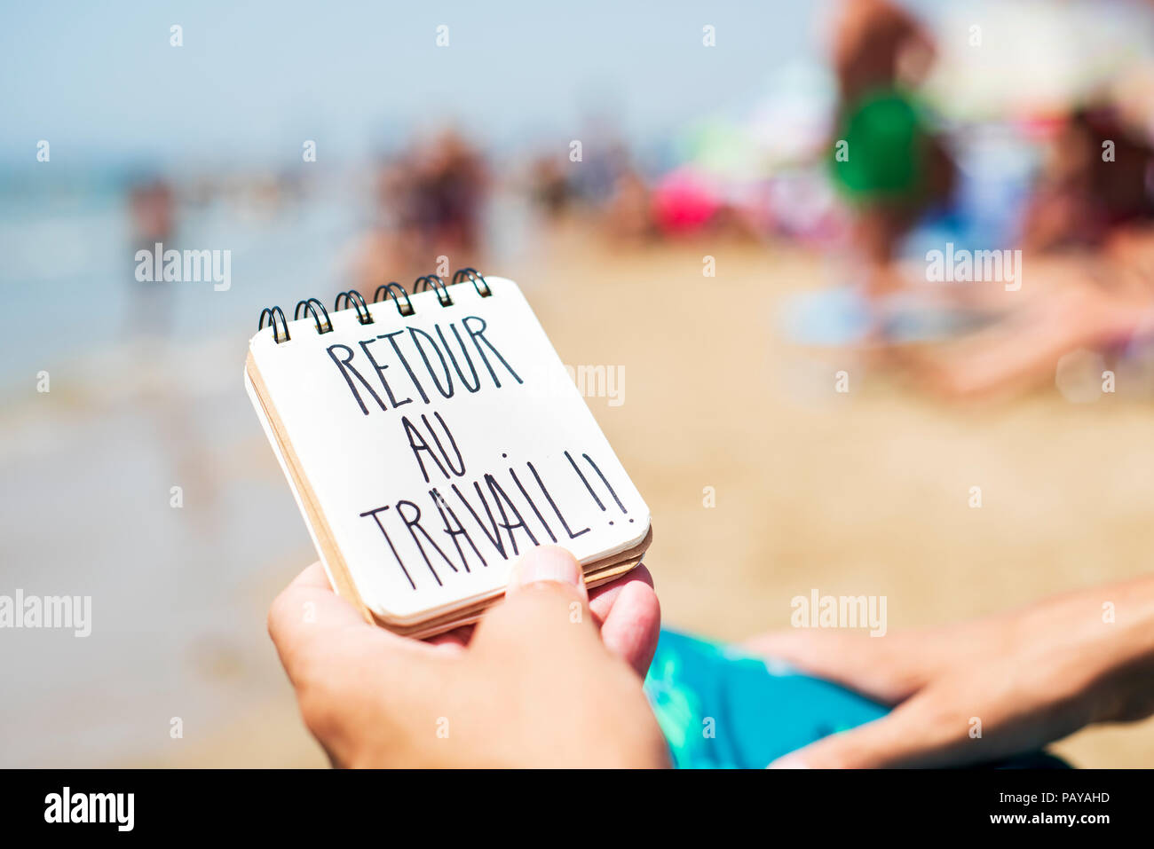 closeup of a young caucasian man sitting in a deck chair in the seashore of a beach showing a spiral notepad with the text retour au travail, back to  Stock Photo