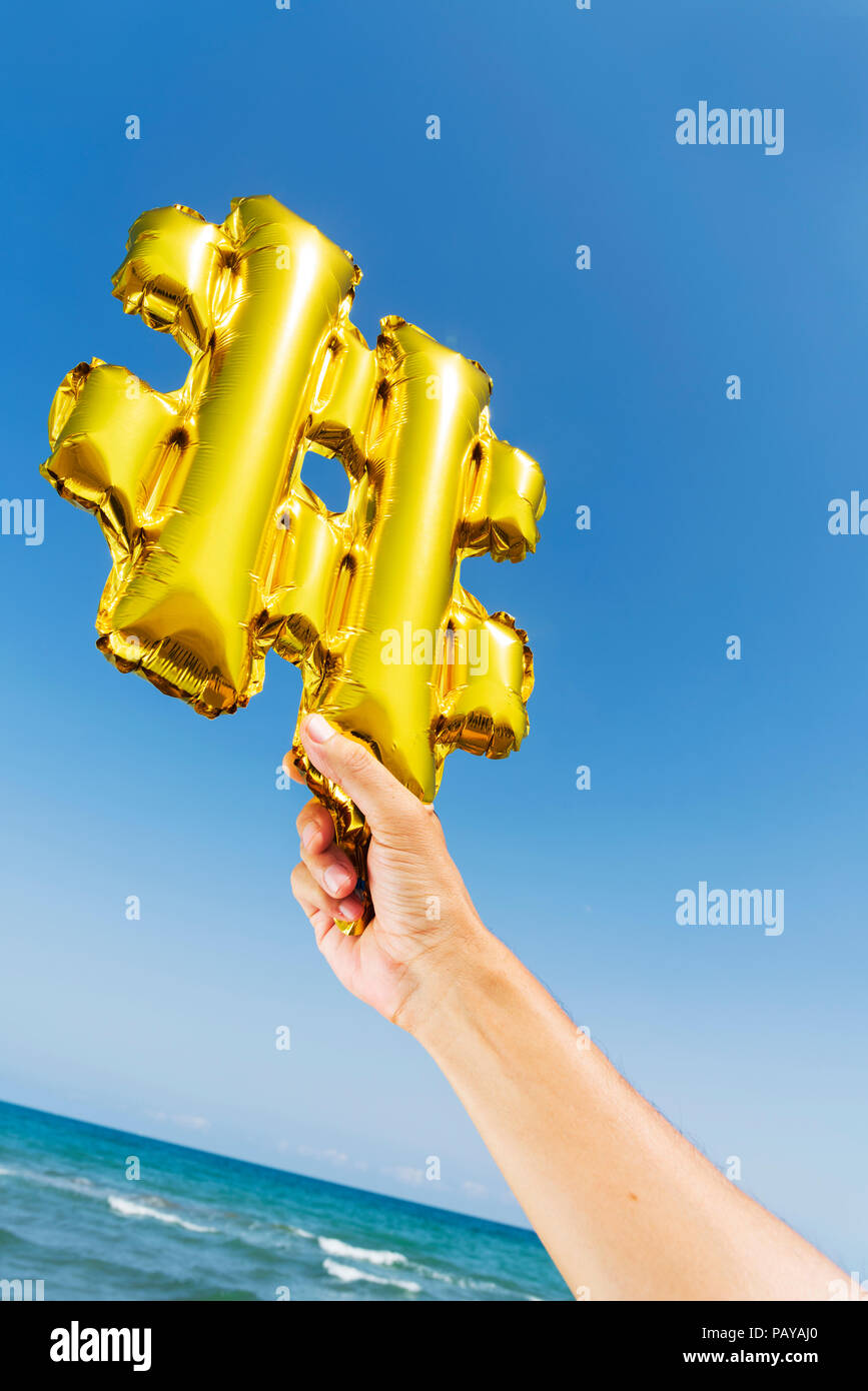 closeup of the hands of a young caucasian man on the beach holding a balloon in the shape of a hash symbol against the sky, with the sea in the backgr Stock Photo