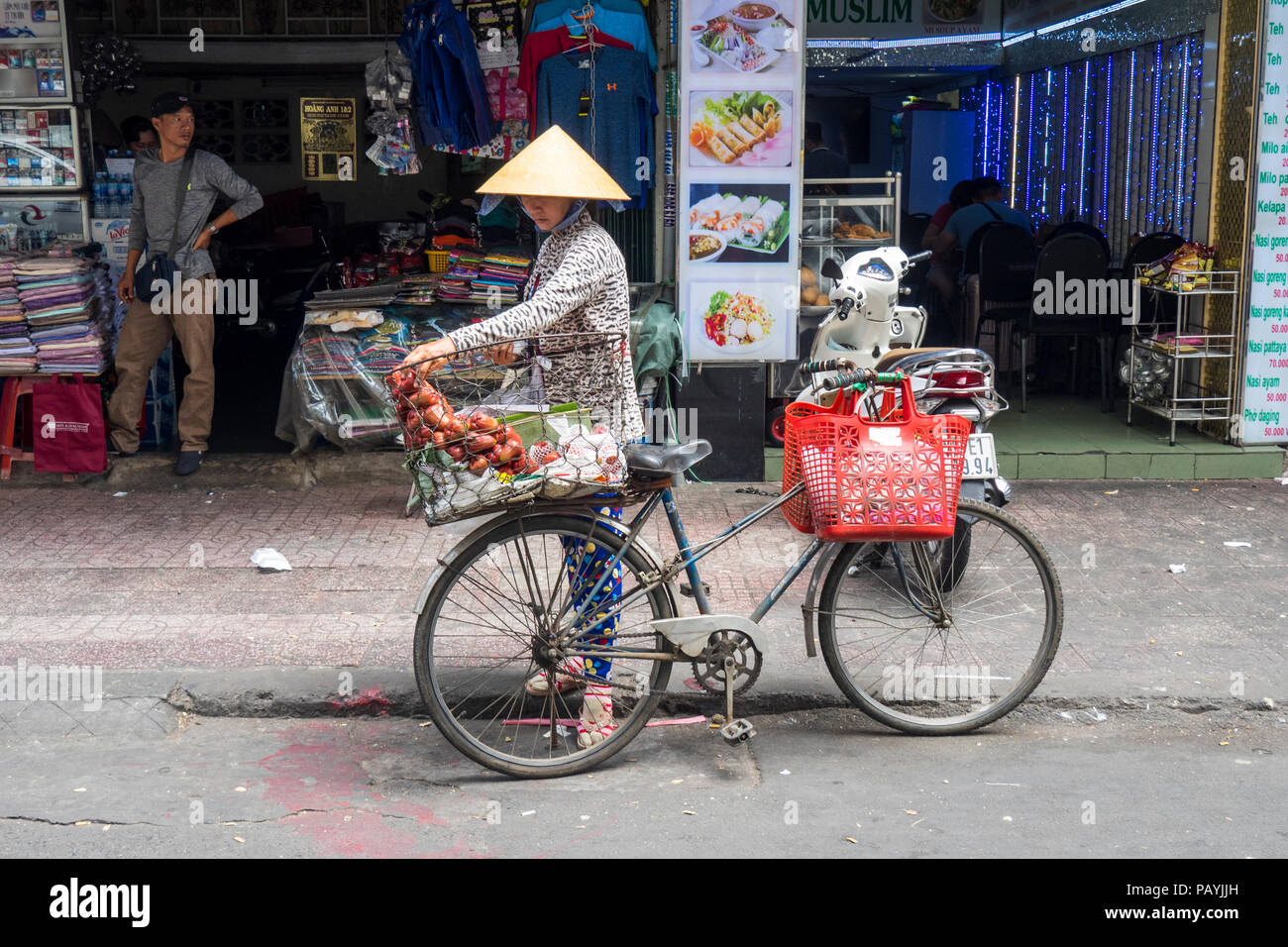 Vietnamese woman wearing a traditional straw conical hat placing her shopping in a basket on her bicycle in Ho Chi Minh City, Vietnam. Stock Photo