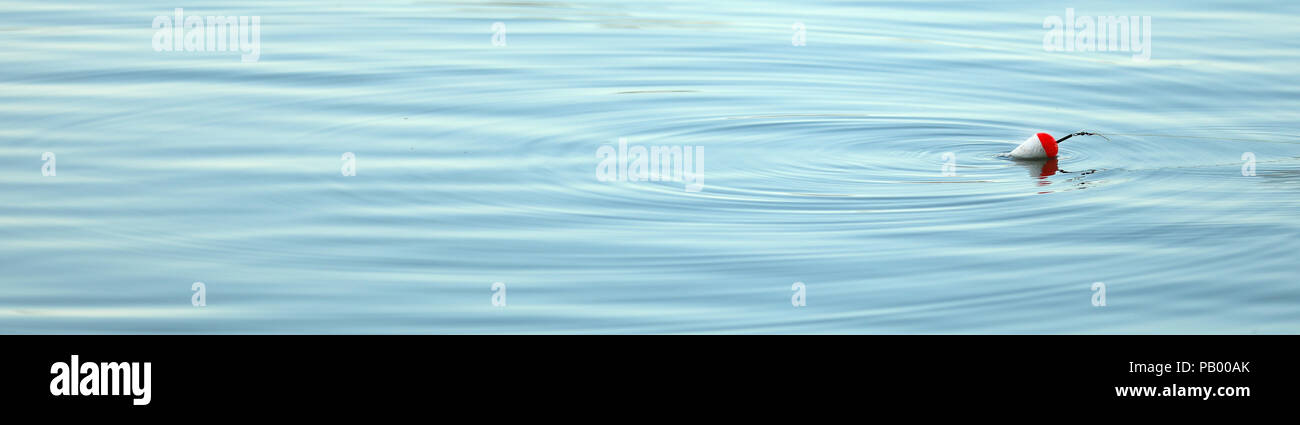 a simple fishing float bobbing on silky smooth cool blue water. Gentle soft pond waves or ripples surround the float. retirement calm peaceful time Stock Photo