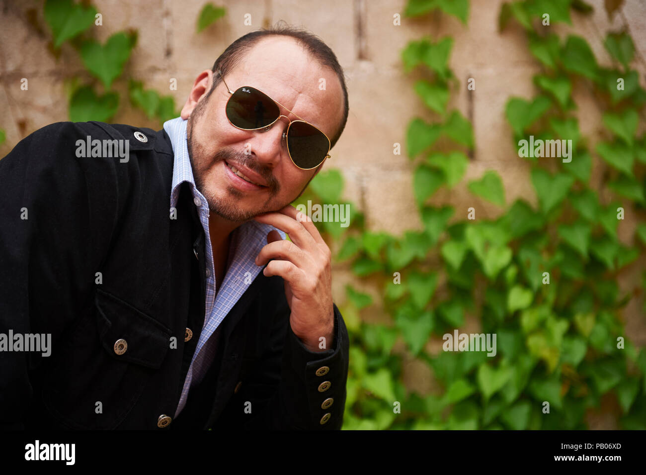 A man poses outdoor, wearing green sunglasses, a blue plaid shirt and a black jacket Stock Photo