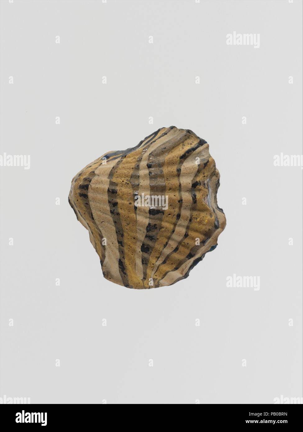 Glass mosaic ornament in the form of a shell. Culture: Roman. Dimensions: H.: 1 3/4 in. (4.4 cm). Date: ca. 1st century A.D..  Translucent cobalt blue, appearing black, with overlays in opaque white and yellow.  Solid block with flat underside in the shape of a bivalve mollusc with radiating ribs.  Decoration of nine marvered trails applied across shell in alternating yellow and white.  Intact, but chipped around edges; dulling, pitting of surface bubbles, and faint iridescence.  This unusual object may have served as part of a wall inlay. Museum: Metropolitan Museum of Art, New York, USA. Stock Photo