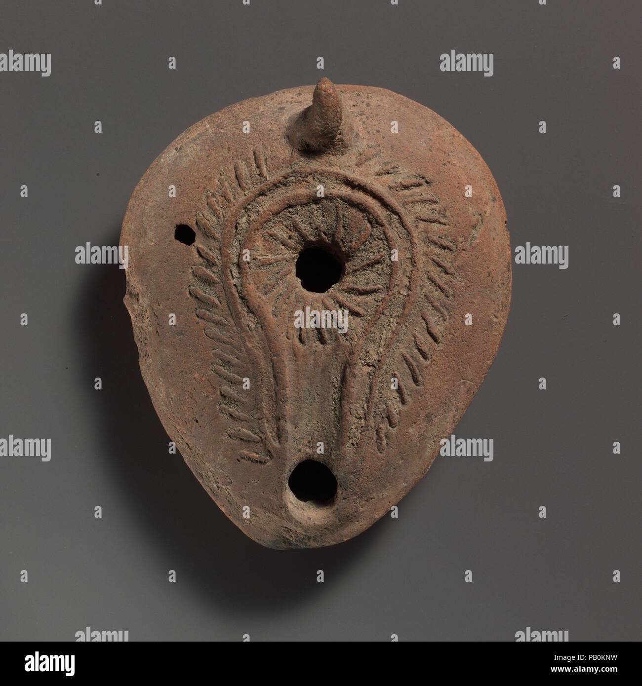 Terracotta oil lamp. Culture: Roman. Dimensions: Overall: 1 1/2 x 3 5/8 in. (3.8 x 9.2 cm). Date: 7th century A.D..  Mold-made, with applied handle. Discus: 'Catherine wheel' lines radiating from a single central filling hole, surrounded by a double ridge; the inner ridge continuing forward around the wick hole, forming a shallow channel. On the broad, sloping shoulder, slanting strokes round discus and nozzle channel. Small, solid lug handle, slightly hooked at rear. sharply carinated body. Circular hollow on base; linear ridge from base to underside of nozzle.  Intact, except for small hole  Stock Photo