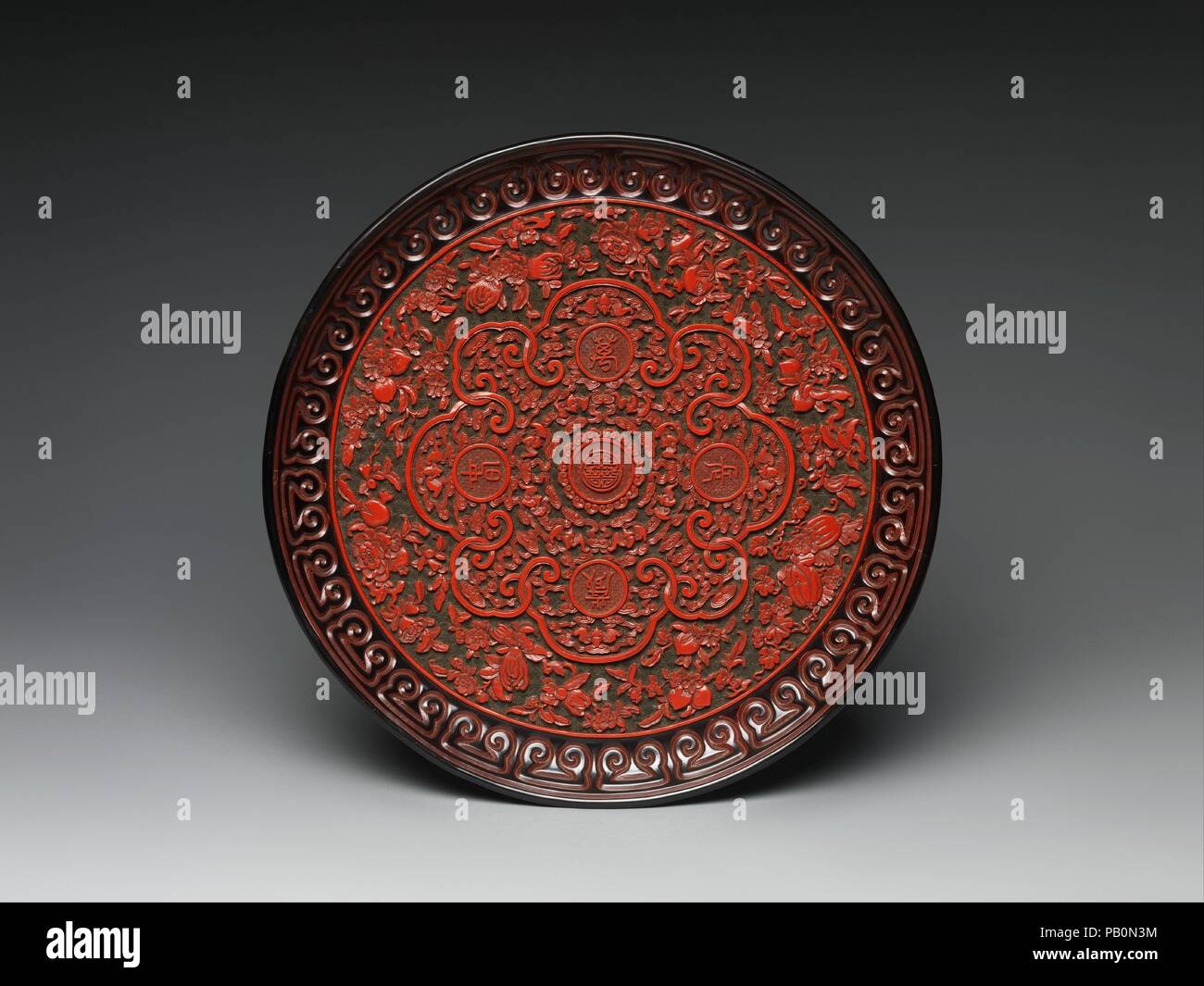 Tray with Chinese characters, bats, and fruits. Culture: China. Dimensions: Diam. 14 1/2 in. (36.8 cm). Date: 1736-1795.  The dense, multilayered surface of this dish is typical of Qianlong-period carved lacquer. At center is the character for longevity (shou) surrounded by first a circle of bats and then a lobed cartouche containing the phrase 'ten thousand longevities and eternal spring' (wan sui chang chun). The outer border of fruits and flowers also refers to long life. The dish was likely presented as a birthday present or used to proffer and display a gift. Museum: Metropolitan Museum o Stock Photo