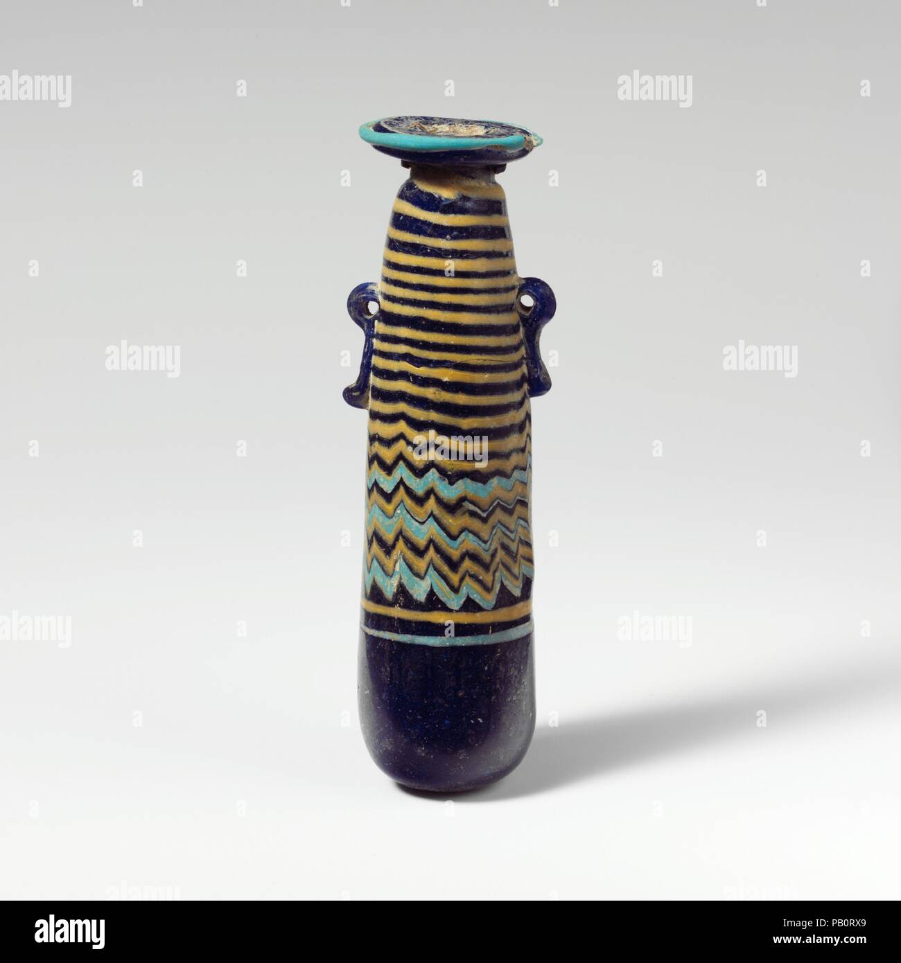 Glass alabastron (perfume bottle). Culture: Greek, Eastern Mediterranean. Dimensions: H.: 4 5/16 in. (11 cm). Date: late 6th-5th century B.C..  Translucent cobalt blue, with handles in same color; trails in opaque yellow and opaque turquoise blue.  Broad, uneven horizontal rim-disk, with slightly raised edge around mouth; short cylindrical neck; narrow rounded shoulder; straight-sided cylindrical body, tapering upwards; convex bottom; two vertical ring handles with knobbed tails, applied over trail decoration.  Turquoise blue trail attached at edge of rim-disk; a yellow trail applied on unders Stock Photo