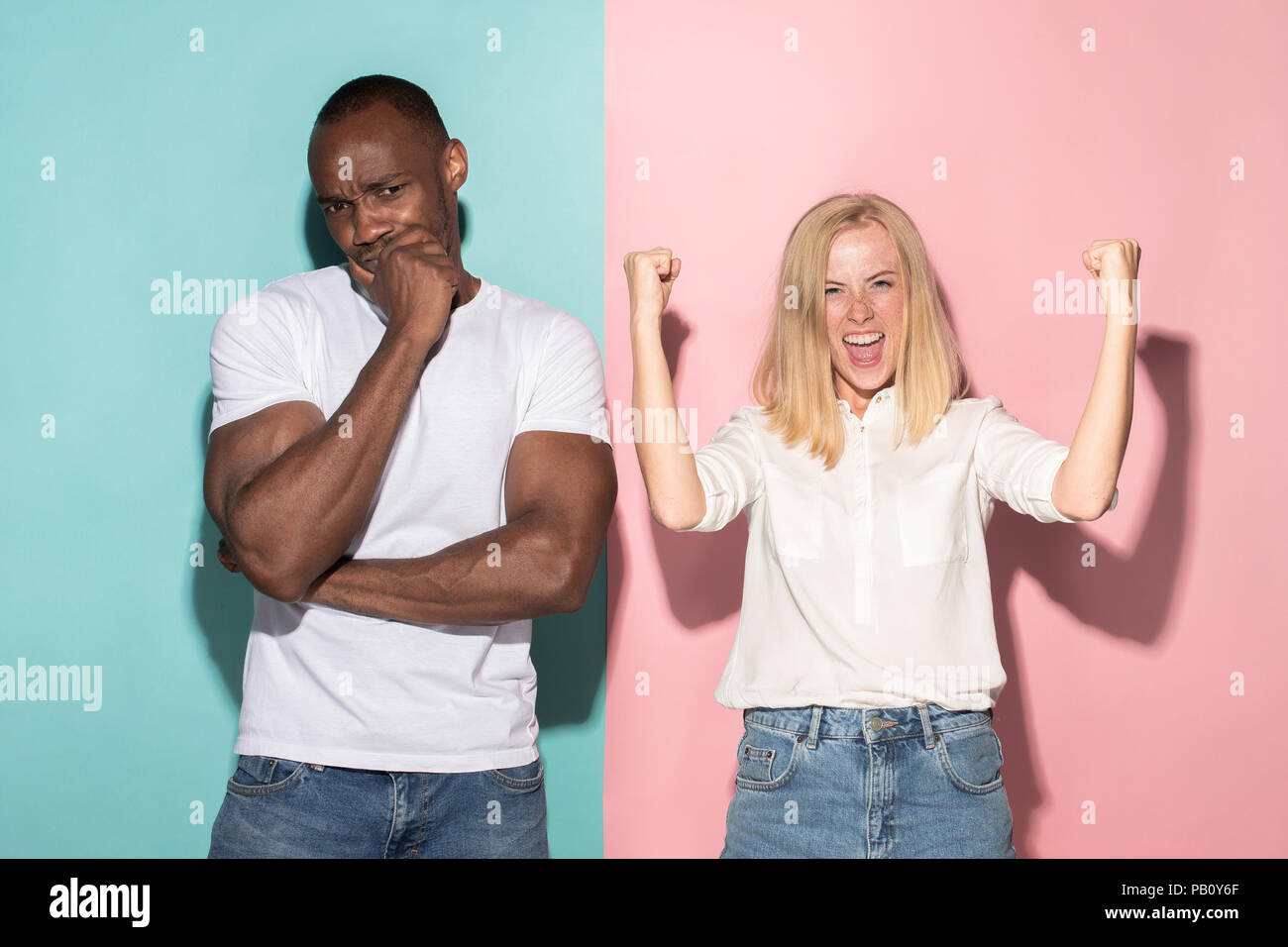 Closeup portrait of young couple, man, woman. One being excited happy smiling, other serious, concerned, unhappy on pink and blue background. Emotion contrasts Stock Photo