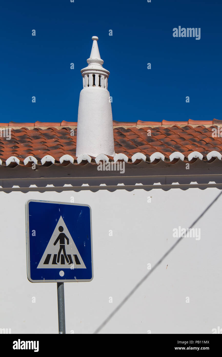 White clean wall of a house with orange roof tiles and typical portuguese chimney. Traffic sign pedestrian crossing. Bright blue sky. Estoi,Portugal. Stock Photo