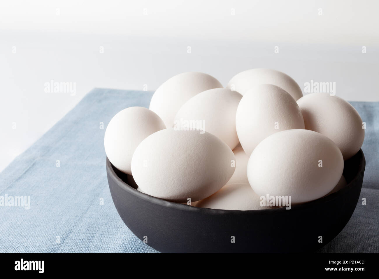 White eggs in a black bowl on blue linen napkin on white background from side off centre composition Stock Photo