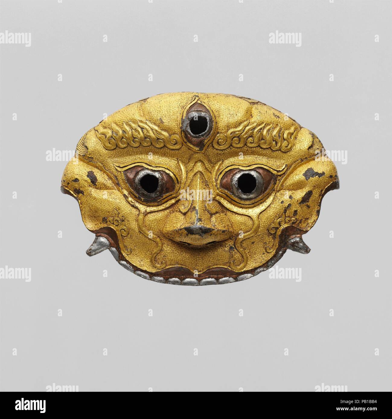 Sword Guard. Culture: Tibetan or Chinese. Dimensions: H. 3 1/4 in. (8.3 cm); W. 4 3/4 in. (12.1 cm). Date: 14th-15th century.  Depicting the face of a wrathful Tibetan Buddhist guardian deity, this extremely rare sword guard was originally part of a complete and very lavish sword. It is from the peak period of Tibetan or Sino-Tibetan ironwork, coinciding with the rule of the Phagmodrupa kings in central Tibet and the Hongwu and Yongle dynasties in China. It is exceptional for the precision and crispness of its chiseling, punched work, and damascening; for the height of the raised decoration; a Stock Photo