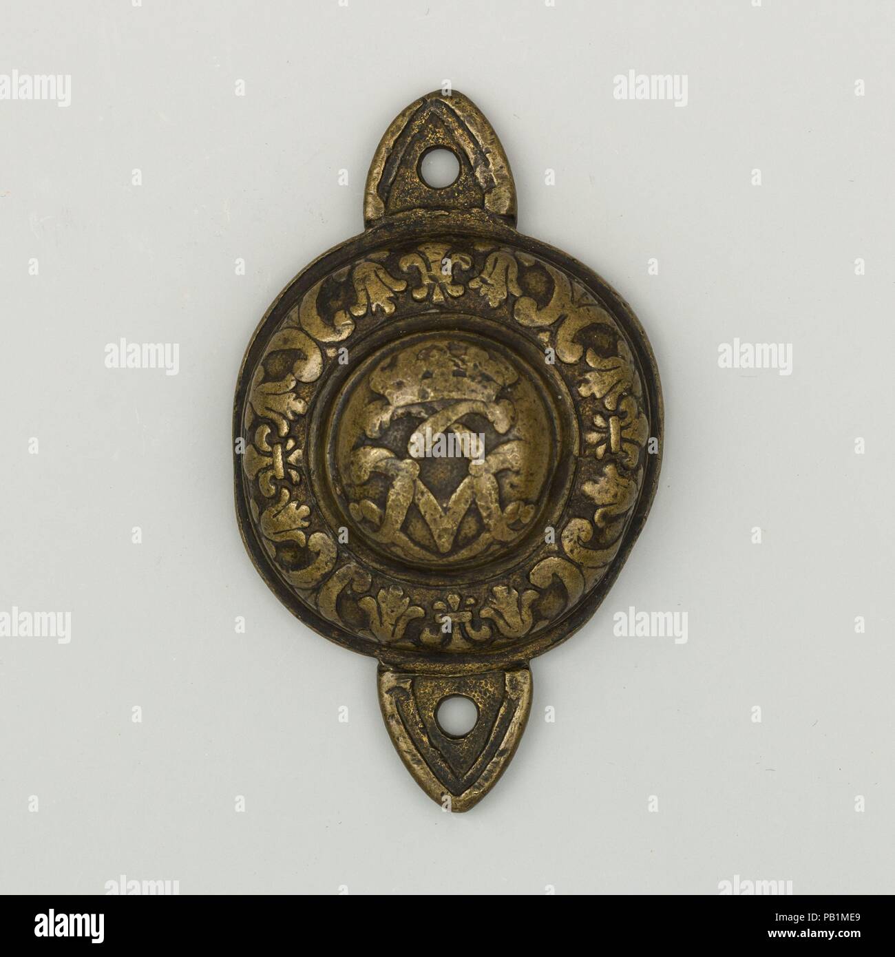 Bit Boss. Culture: French. Dimensions: Diam. 3 5/16 in. (8.4 cm); Wt. 2.4 oz. (68 g). Date: first half 18th century.  The rim of this bit boss is decorated with four fleurs-de-lis. At its center are the two interlaced Ls of King Louis XIV (r. 1643-1715) or, more likely, King Louis XV (r. 1715-1774), crowned at the top. What looks like a letter V behind the monogram is actually a very simplified representation of the two crossed palms often seen on this kind of composition.  This symbolism suggests that this boss decorated the side of a bit worn by a cavalry or a coach horse used by the French  Stock Photo