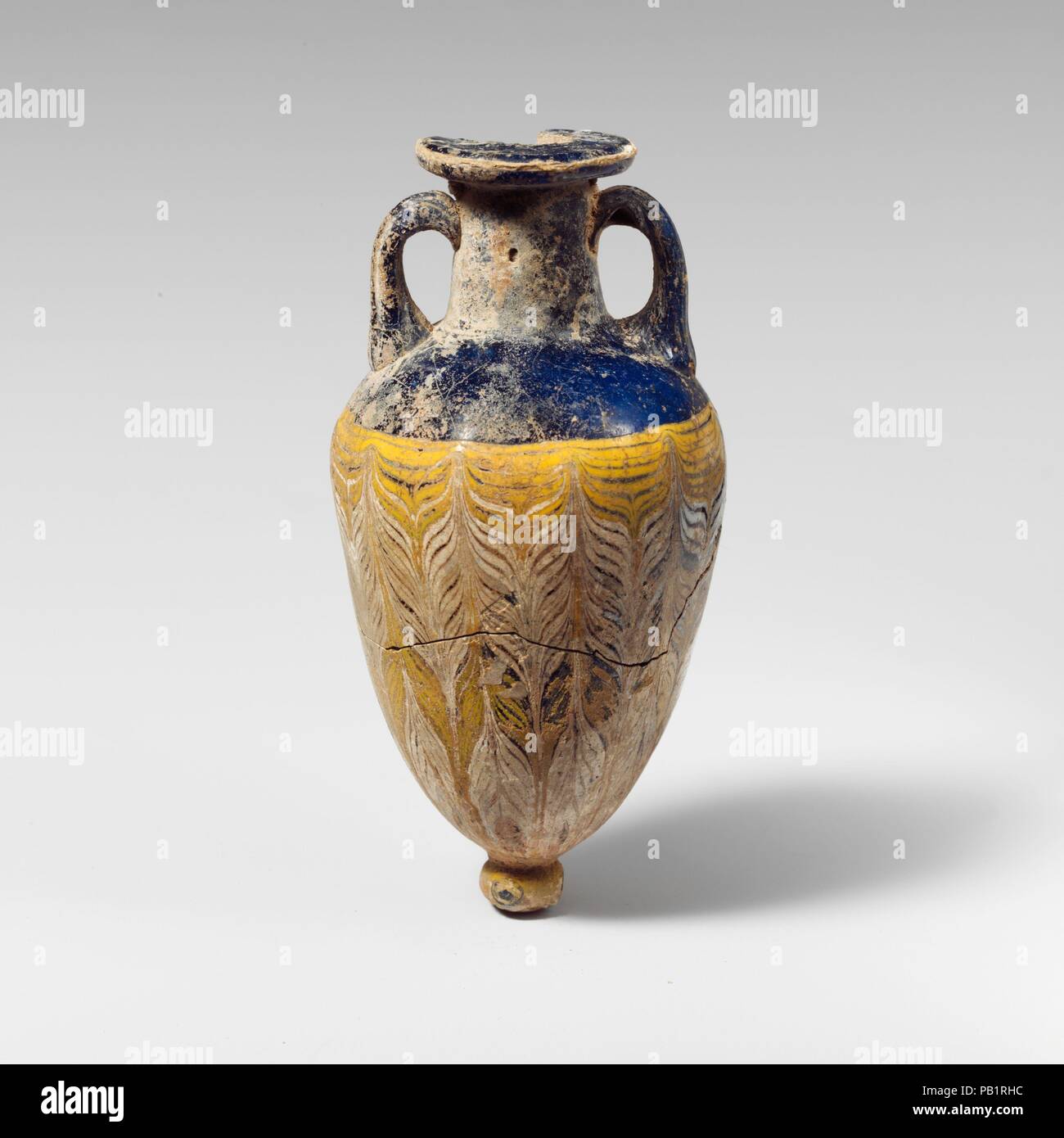 Glass amphoriskos (perfume bottle). Culture: Greek, Eastern Mediterranean or Italian. Dimensions: H.: 4 3/8 in. (11.1 cm). Date: 4th-3rd century B.C..  Translucent cobalt blue, with handles and base-knob in same color; trails in opaque yellow and opaque white.  Broad horizontal rim-disk with rounded edge; cylindrical neck, expanding slightly downwards; broad, sloping shoulder; elongated ovoid body, tapering sharply downwards; applied small circular base-knob with uneven bottom; two strap handles applied to shoulder and drawn up, turned in, and pressed on to neck.  One fine white trail attached Stock Photo