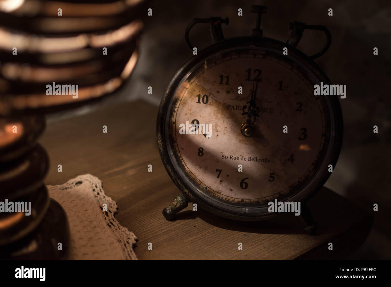 An old clock on a night stand lit partly by a night lamp with thatched hood, creating a calm an romantic mood. An address in Paris, Europe, is on the  Stock Photo