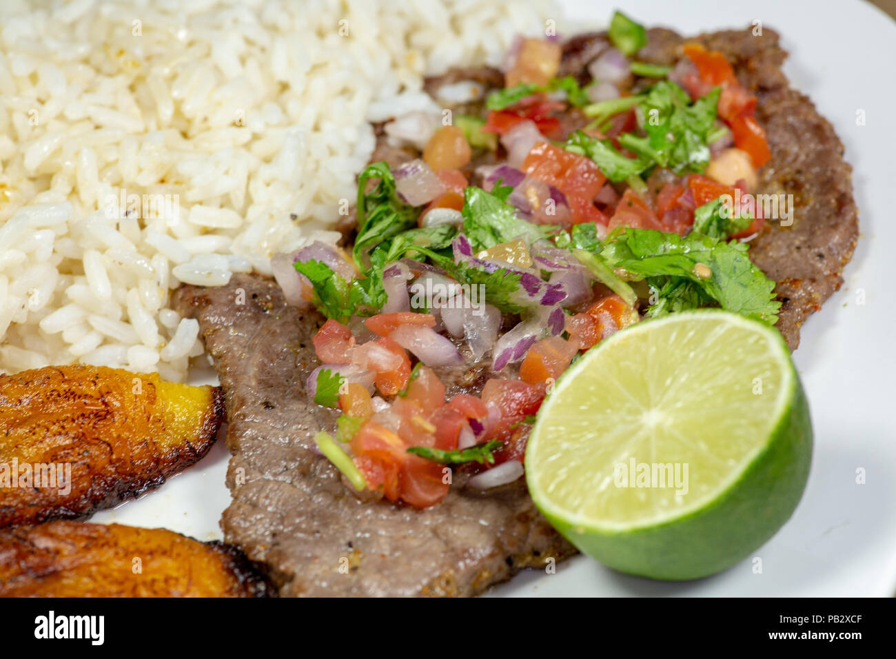 A steak covered in pico de gallo surrounded by plantains and white rice on a white plate. Cuban food. Stock Photo