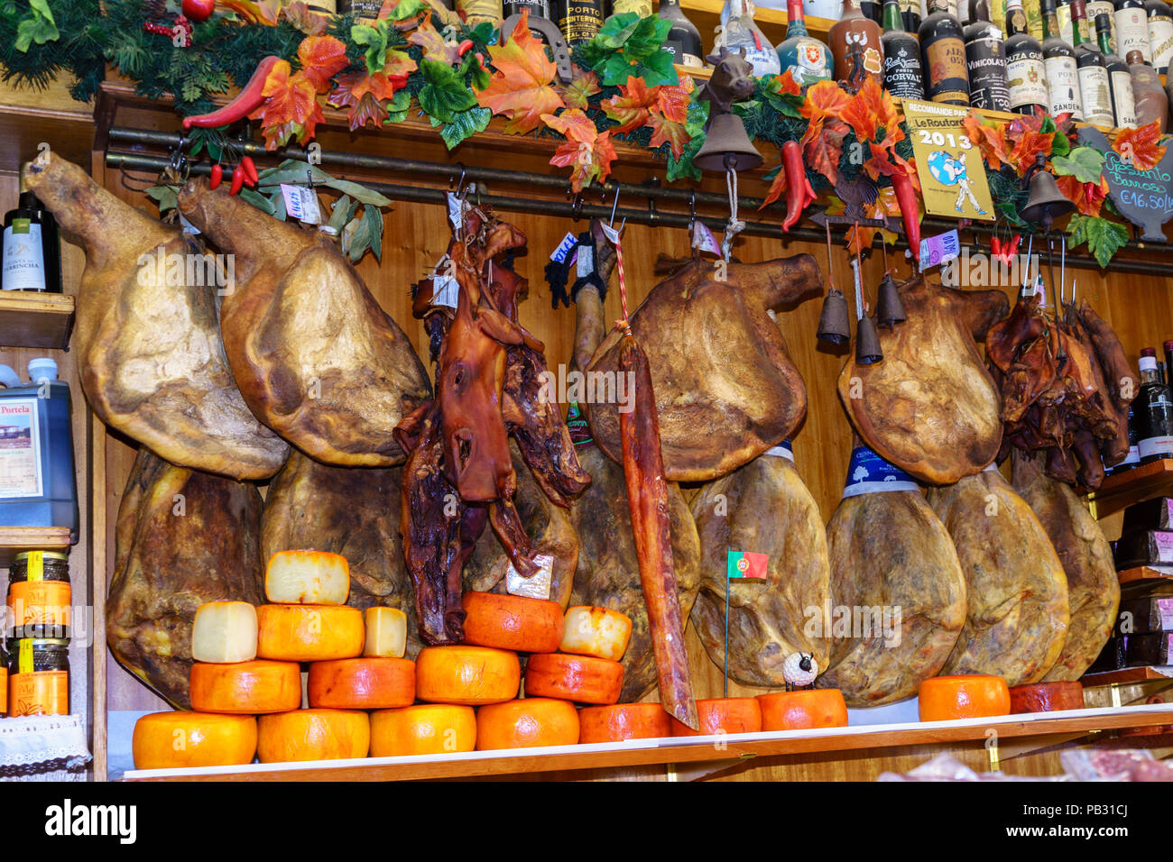 Iberian Ham hanging from hooks in a traditional delicatessan in Portugal. A row of round cheeses are arranged in the front. Stock Photo