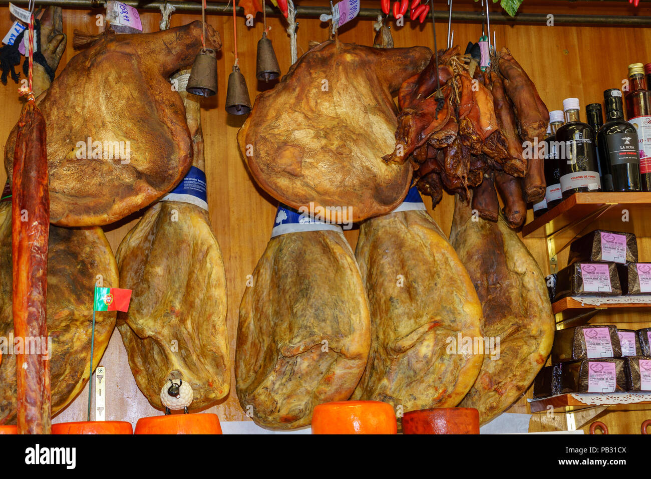 Iberian Ham hanging from hooks in a traditional delicatessan in Portugal. A row of round cheeses are arranged in the front. Stock Photo